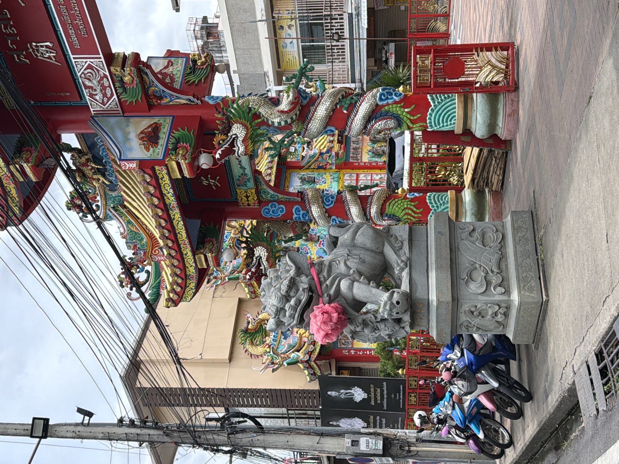 This is a vertical, full-shot, eye-level, outdoor photograph taken during the day. The image depicts a brightly colored, ornate Chinese-style archway or temple entrance with intricate carvings of dragons and other mythical creatures. The structure is predominantly red with gold and green accents, featuring detailed paintings and sculptures.
In the foreground, to the right, several motorcycles are parked along a paved walkway. The motorcycles are in various colors including blue, white, and pink. A person, wearing a helmet, is partially visible near the motorcycles.
Behind the archway, there are buildings and an overcast sky. The environment appears to be urban, with evidence of utility poles and tangled electrical wires in the lower left quadrant of the image, suggesting a busy street or marketplace.
Notable details include the vibrant decorations on the structure, the presence of the parked motorcycles indicating potential foot traffic or a place of worship, and the overhead electrical infrastructure which is common in many parts of Southeast Asia. The weather appears to be cloudy.
There is visible text on signs and the archway, primarily in a language that appears to be Thai or Chinese. One sign behind the statue, in the lower middle, shows black and white portraits with text.
The scene captures a cultural landmark in an urban setting, likely a place of religious significance or a decorative gateway, with everyday elements like parked vehicles and utility infrastructure juxtaposed against its elaborate design.