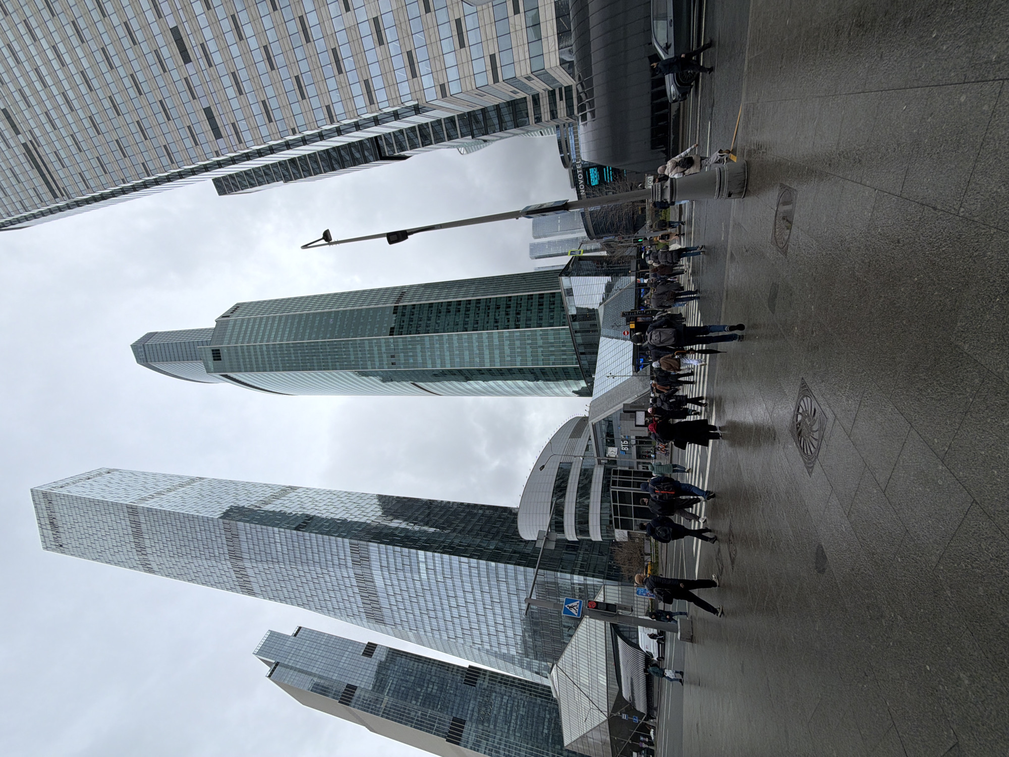 A low-angle, wide shot captures a modern urban street scene dominated by towering skyscrapers under an overcast sky. The image is taken from ground level, looking up at the surrounding architecture.
Numerous pedestrians are visible on the wet, paved sidewalks and crosswalks. They are dressed in various casual and semi-formal attire, walking or standing, suggesting a typical busy city environment. A dark car is partially visible on the far right, and another white vehicle is seen further down the street.
The most prominent features are the multiple high-rise buildings, mostly clad in glass and reflecting the gray sky. One particularly tall, slender skyscraper stands centrally, with another equally tall building to its left. A distinct, modern building with a curved, white roofline is visible towards the right side of the frame, further back. Street lamps, traffic light poles with pedestrian signals, and a trash bin are scattered along the pedestrian areas. The ground appears damp, indicating recent rain or high humidity.
Visible text includes "NOVOTEL" on a building, likely indicating a hotel, and "ВТБ" (VTB), a Russian bank, on another structure. The sky is uniformly gray, suggesting an overcast day without direct sunlight. The scene depicts a bustling city street in Khimki, Russia, characterized by contemporary architecture and active pedestrian life.