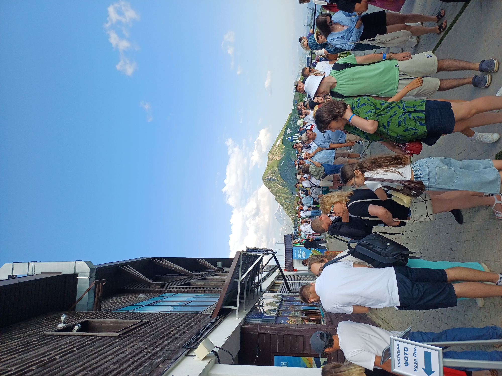 This is a medium-shot, eye-level, outdoor photograph taken on a sunny day in Estosadok, Russia, likely at a mountain resort. The image captures a queue of people waiting outside a wooden building with large windows. In the foreground, several individuals are visible, including a man in a white t-shirt and navy shorts, a woman in a black t-shirt and shorts, and a young girl in denim shorts and a white t-shirt. A sign with Russian text and an arrow pointing left is positioned on the left side of the frame, indicating a photo pickup location named "Rosa Pik." In the background, a large crowd of people are visible, and further beyond, a majestic mountain with a green peak and patches of snow dominates the landscape under a clear blue sky with scattered white clouds. The scene suggests a popular tourist attraction, possibly a ski lift station or a viewpoint.