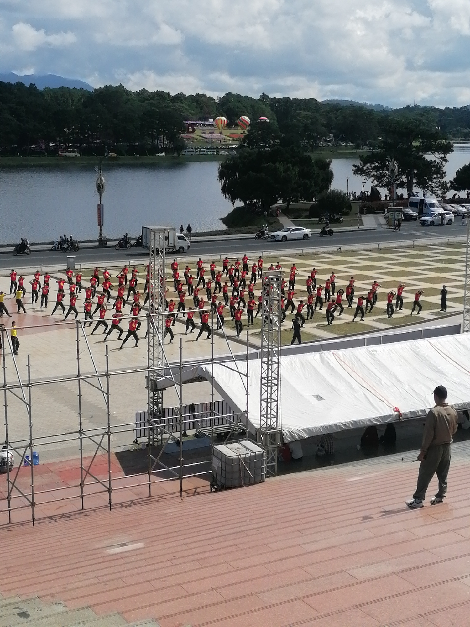 In Da Lat, Vietnam, a large group of people in red shirts and black pants are performing an outdoor dance routine on a paved plaza. The plaza is bordered by a lake to the left and a street with vehicles and pedestrians to the background. A white tent structure, likely for an event, is set up on the right side of the plaza, and scaffolding is visible in the foreground. A man in a tan uniform stands on a red staircase overlooking the scene. In the distance, hot air balloons are visible in the sky above a treeline.