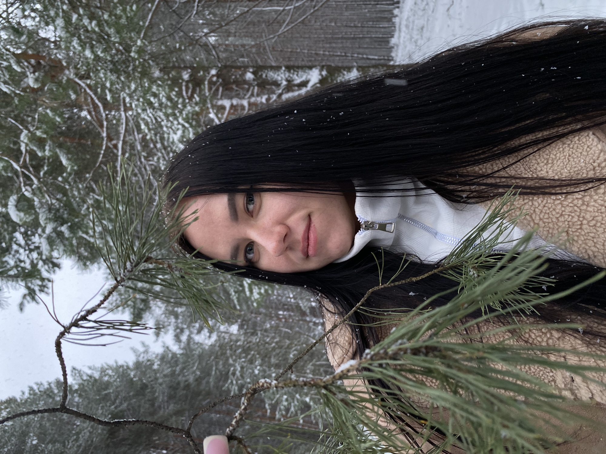 The image shows a woman with long, dark hair posing with a pine branch in a snowy forest. The woman is wearing a light-colored jacket with a zipper. Snowflakes are visible in her hair and on the pine needles. The background features snow-covered trees. The location is Chaikovsky, Russia, as specified.