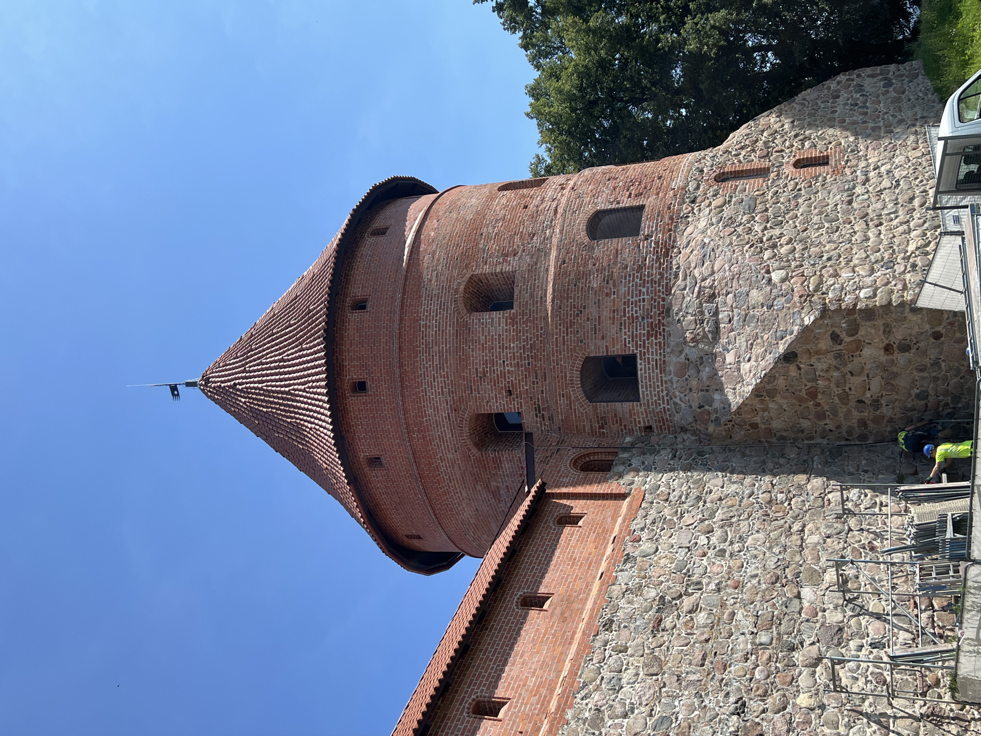 The image shows a close-up, low-angle view of the Trakai Island Castle in Trakai, Lithuania. The focus is on a circular brick tower with a conical red-tiled roof, topped with a metal spire flying a dark flag. The tower is constructed from red brick and features several arched windows, some of which appear dark. The lower portion of the tower and the adjacent wall are made of rough-hewn stones. To the left of the tower, a section of a brick wall with a red-tiled roof runs horizontally, also constructed with stone at its base and featuring small rectangular windows. The wall is undergoing some form of construction or renovation, with scaffolding and construction materials visible at its base. Two individuals, dressed in high-visibility safety vests and hard hats, are working near the scaffolding. One person, wearing a yellow vest and blue helmet, is standing and appears to be working on the scaffolding. Another person, wearing a bright yellow vest, is bent over near the ground. In the background, to the right of the tower, is a dense canopy of green trees. The sky is clear and bright blue.