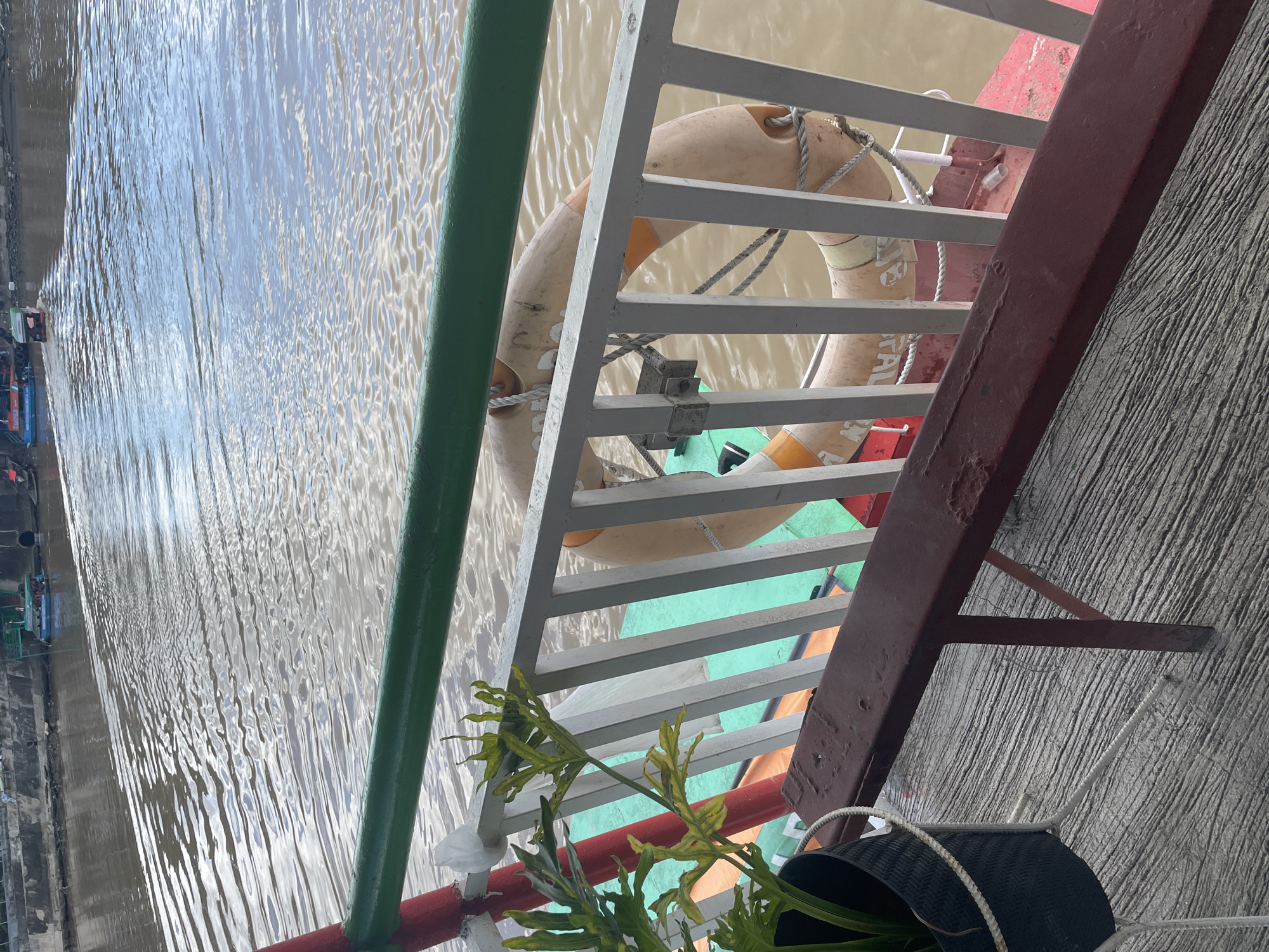 The image shows a view from a boat on a river in Davao City, Philippines. In the foreground, there is a wooden bench with a metal frame, a black planter with a green leafy plant, and a white railing. A life preserver is attached to the railing. In the background, the river flows with small boats and a concrete embankment visible on the far bank. The water is muddy brown, and the sky is overcast. The overall scene depicts a calm, somewhat weathered, outdoor environment on a body of water.