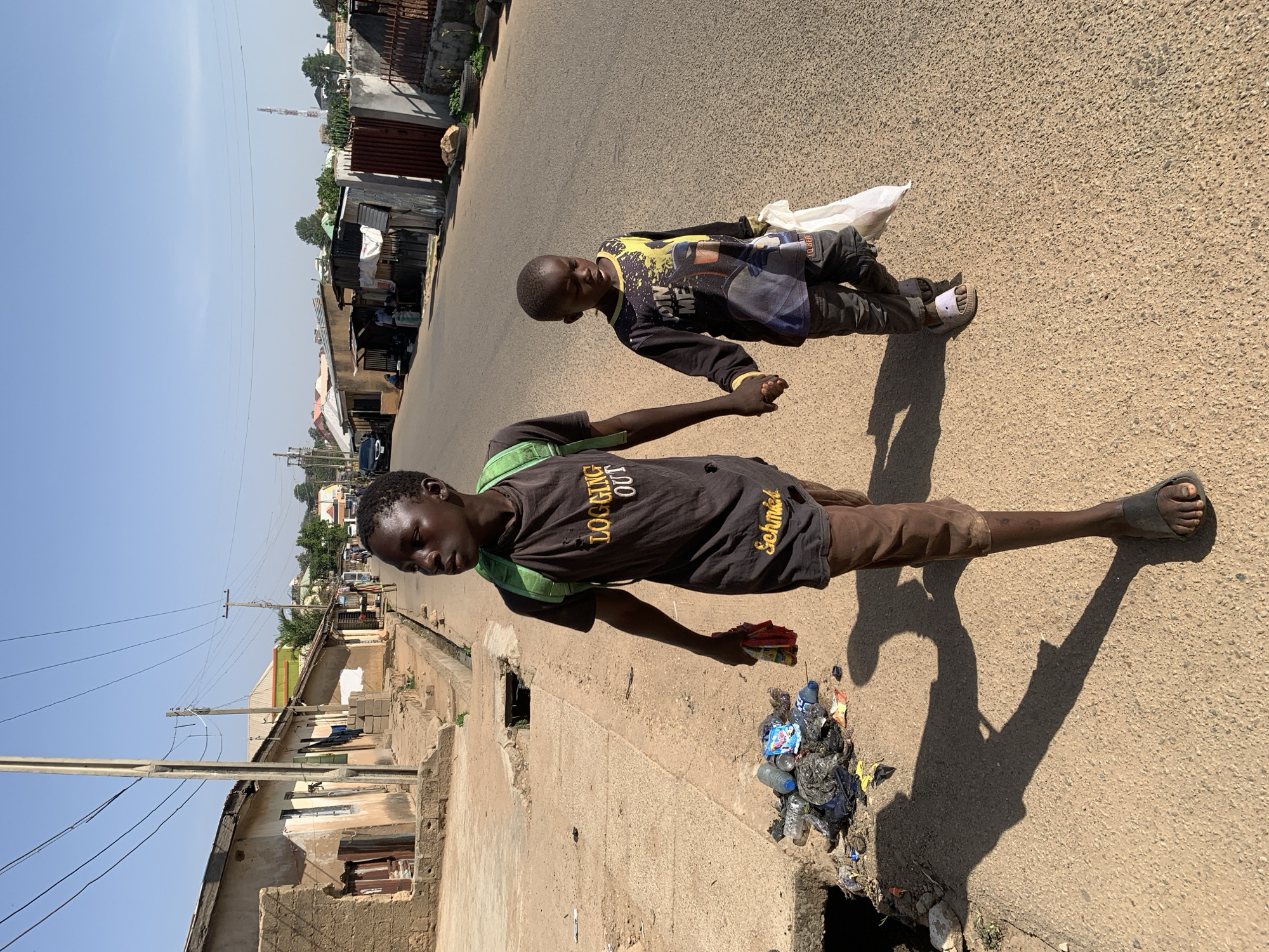 Two young boys are walking along a paved street in Jos, Nigeria. The older boy wears a black shirt, brown shorts, and sandals, and carries a green backpack. He is holding a small packet in his left hand. Next to him, in the street gutter, is a pile of plastic bottles and other trash. The younger boy wears a long-sleeved shirt, pants, and sandals, and carries a white plastic bag. The two boys hold hands as they walk. The background shows a mix of residential and commercial buildings, some of which appear to be in a state of disrepair. Power lines are visible in the sky above the buildings.