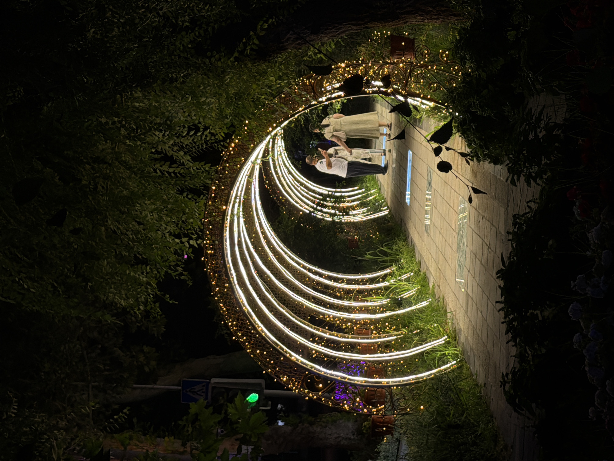 The image depicts a nighttime scene on a paved walkway in Chirchik, Uzbekistan, illuminated by a decorative archway of white lights and numerous small, warm-toned string lights. Three individuals are visible within the archway. On the left, a person wearing a white t-shirt and dark pants stands facing away from the camera, holding a smartphone. To their right, a person in a white outfit, possibly a child, stands facing the same direction. Further to the right, a person in a long, light-colored dress stands with their back to the camera, holding a bag in their left hand. The walkway is flanked by lush greenery and foliage. A street sign, partially obscured by trees, is visible on the left side of the image, indicating a pedestrian crossing. The overall atmosphere is festive and inviting, suggesting a public space adorned for an evening event or attraction.