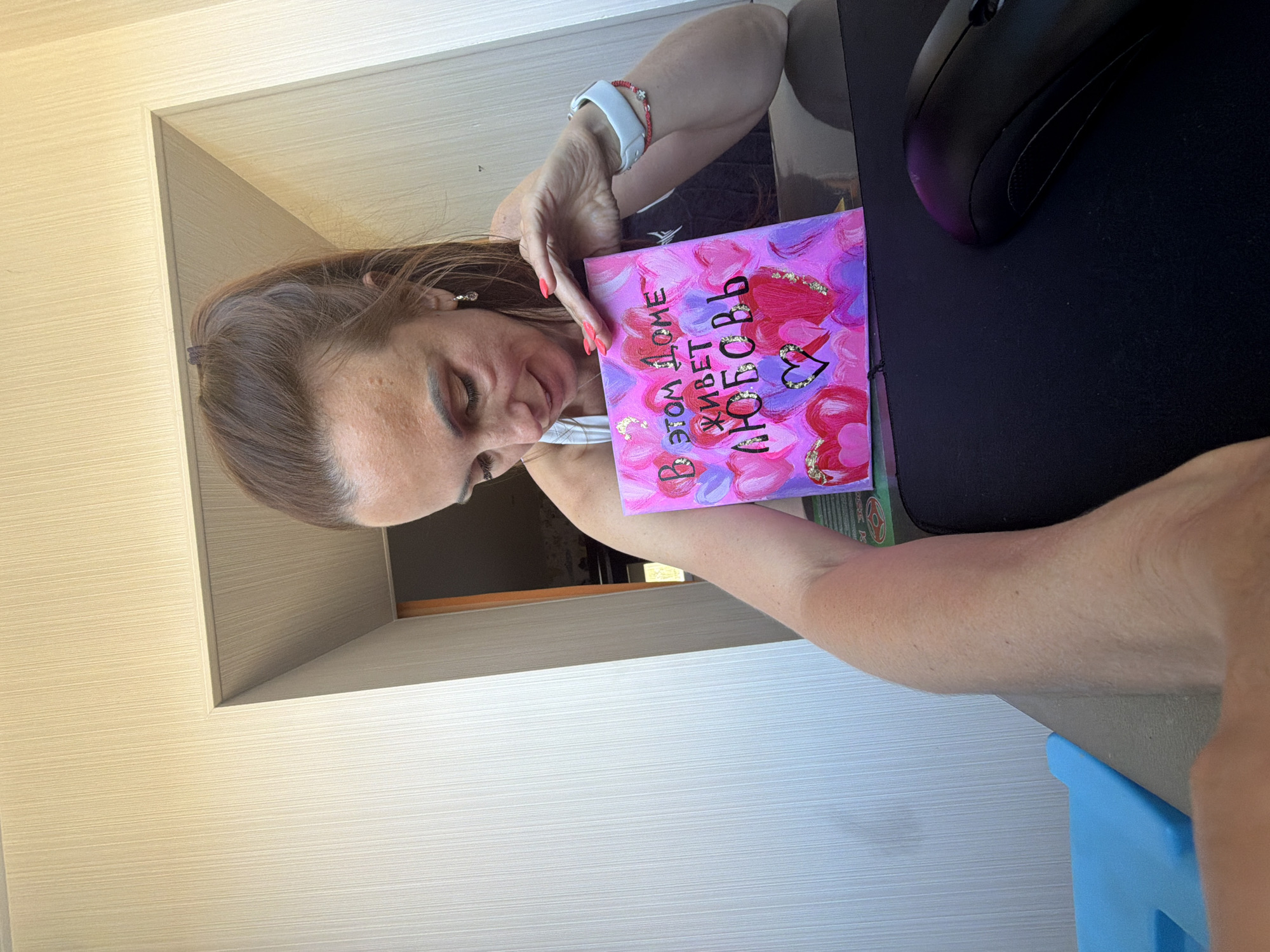 A woman with brown hair pulled back in a ponytail is sitting at a desk, looking down at a small, heart-adorned canvas. The canvas has pink and purple paint with gold accents and reads "В этом доме живет любовь" which translates to "Love lives in this house." She is wearing a white tank top and has a white smartwatch on her left wrist, as well as a red beaded bracelet. On the desk, a black computer mouse is visible. The background shows a doorway with wood-paneled walls and a textured wallpaper on the left. The scene appears to be indoors, possibly in a home office. There is no specific indication of the location being Birobidzhan, Russia.