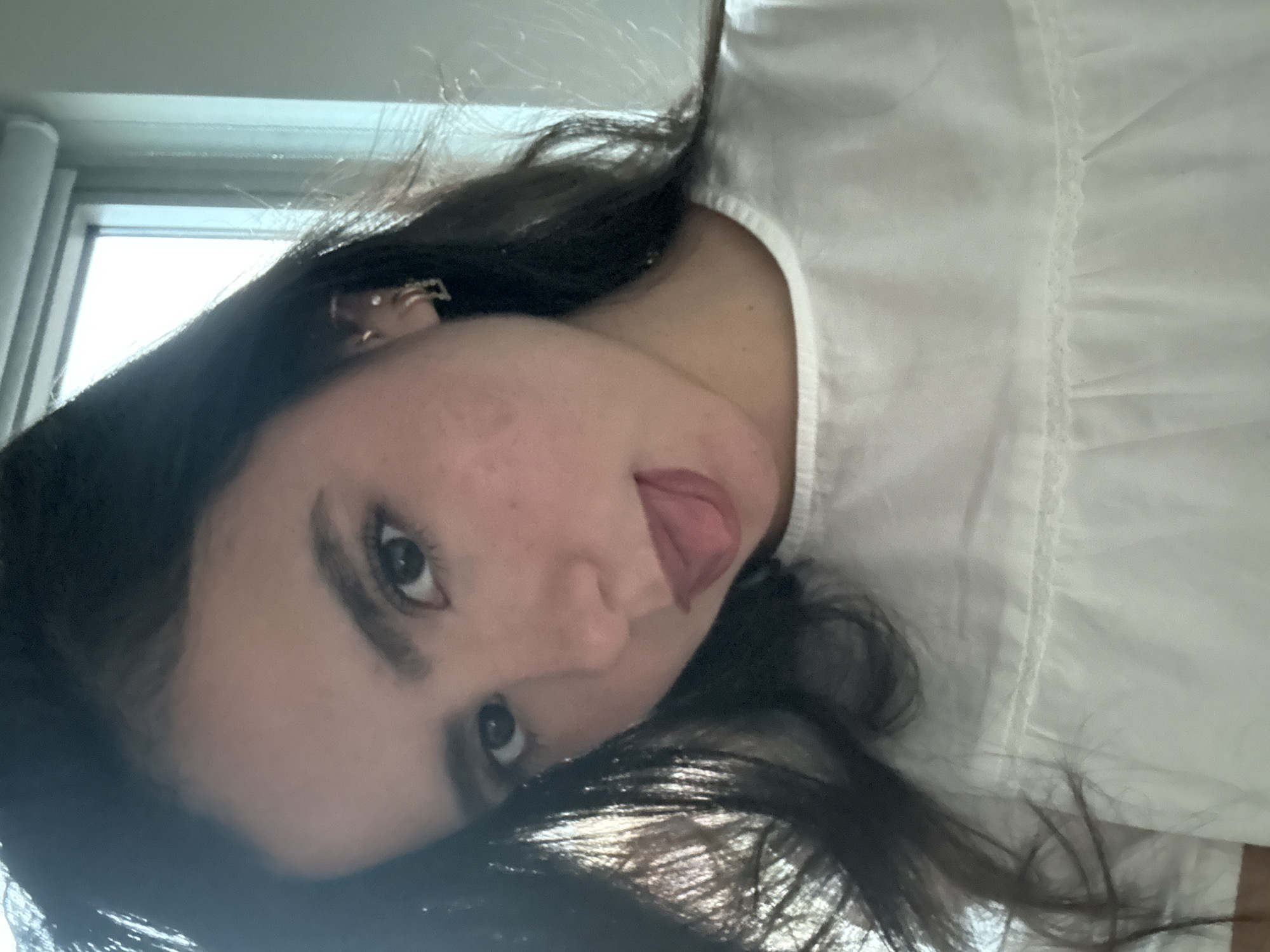 A young woman with dark hair and brown eyes is pictured in a selfie. She is wearing a white top with a ruffled neckline and has her tongue sticking out slightly. She is also wearing a small gold earring in her left ear. The background is blurry and appears to be an indoor setting with a window. The image is taken from the chest up, with her face as the primary focus. The lighting suggests it is daytime, possibly near a window with natural light. No specific location in Austin, United States of America, can be determined from the image.