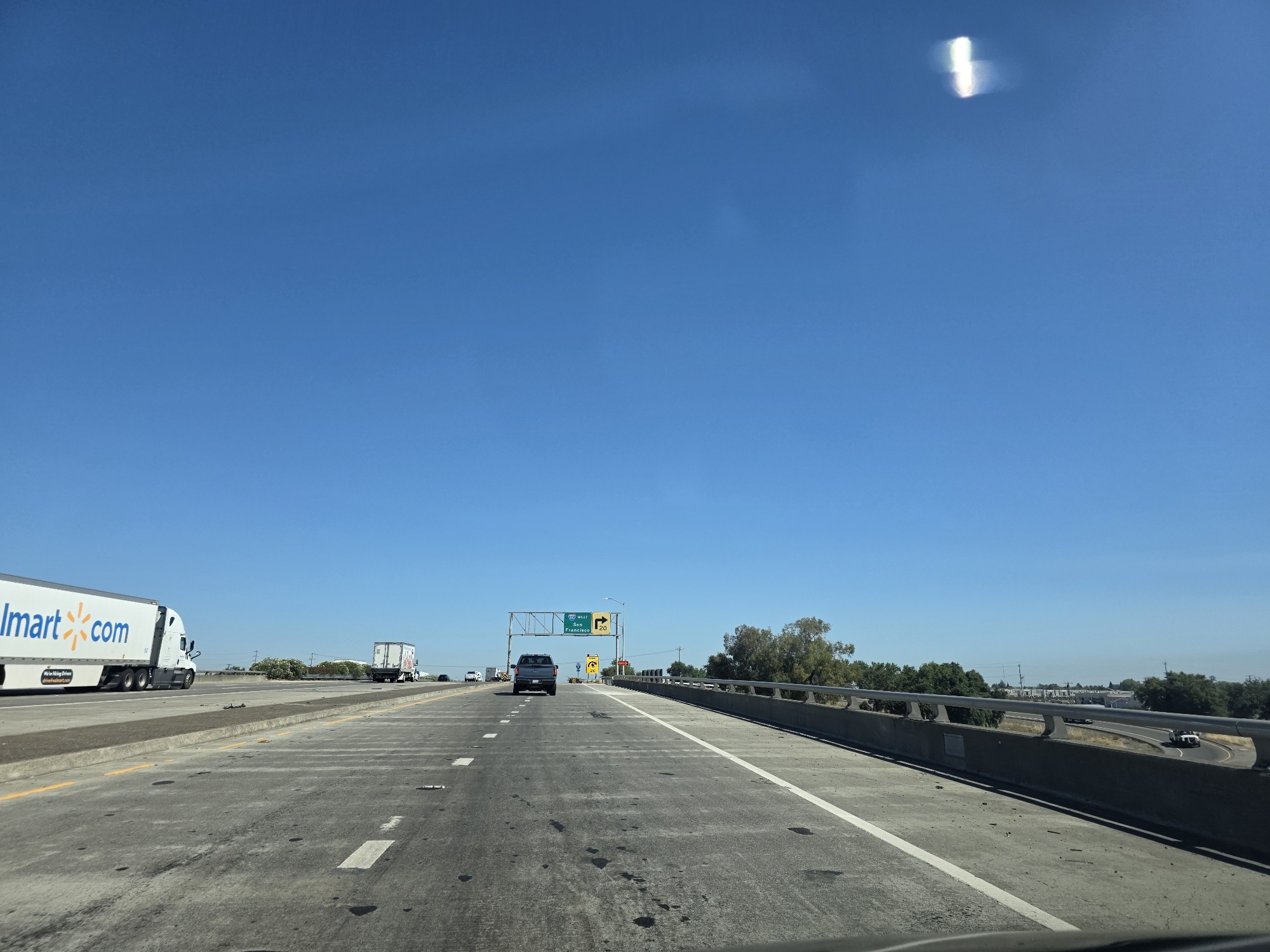 This media file depicts a view from a vehicle traveling on a highway. The scene is set in Sacramento, United States of America, under a clear blue sky, suggesting a sunny day. The highway has multiple lanes marked with white lines, and a concrete barrier runs along the right side.
In the foreground, the road surface shows some dark stains. In the middle ground, several vehicles are visible: a Walmart truck with "Walmart.com" branding is on the left, a dark-colored SUV is in the center lane, and other trucks and cars are further down the road. Overhead, a highway sign indicates "I-80 West San Francisco" and "20" with an arrow pointing to the right, suggesting an upcoming exit.
In the background, there are trees and some buildings, hinting at the transition from rural to urban or suburban landscape. The bright sunlight and clear sky indicate it is daytime, likely late morning or early afternoon. There is a bright, elongated light streak in the upper right portion of the sky, possibly a lens flare or reflection. The overall impression is one of normal highway travel on a pleasant day.