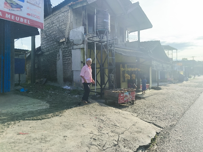 The image shows a street scene in Cikalong, Indonesia (latitude: -6.761, longitude: 107.447). A man in a pink shirt and white cap walks along a sidewalk in front of a row of brick buildings. The buildings are lined with shops and businesses, including a "Mebel" (furniture) store and a stall selling snacks. There is a water tank on a metal frame in front of one of the buildings. A woman in a long blue dress walks in the distance, and the street is paved with grey asphalt. The scene is bathed in sunlight, with a hazy atmosphere.