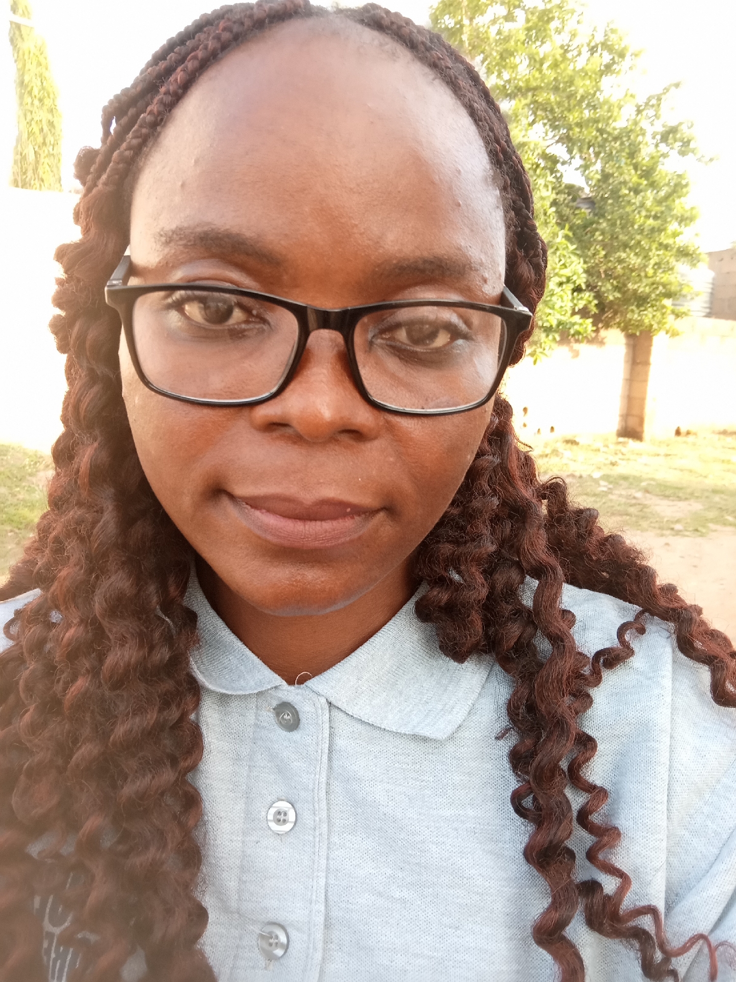 This is a medium close-up, selfie-style photograph of a Black woman wearing glasses and a light blue collared shirt. She has dark, braided hair that extends past her shoulders in curls. Her expression is neutral, with a slight hint of a smile. The background is outdoors, with a blurred tree and a building visible. The lighting suggests it is daytime, possibly late afternoon given the warm tones. The setting appears to be a residential or semi-rural area, consistent with the provided location context of Jalingo, Nigeria. There is no visible text in the image.