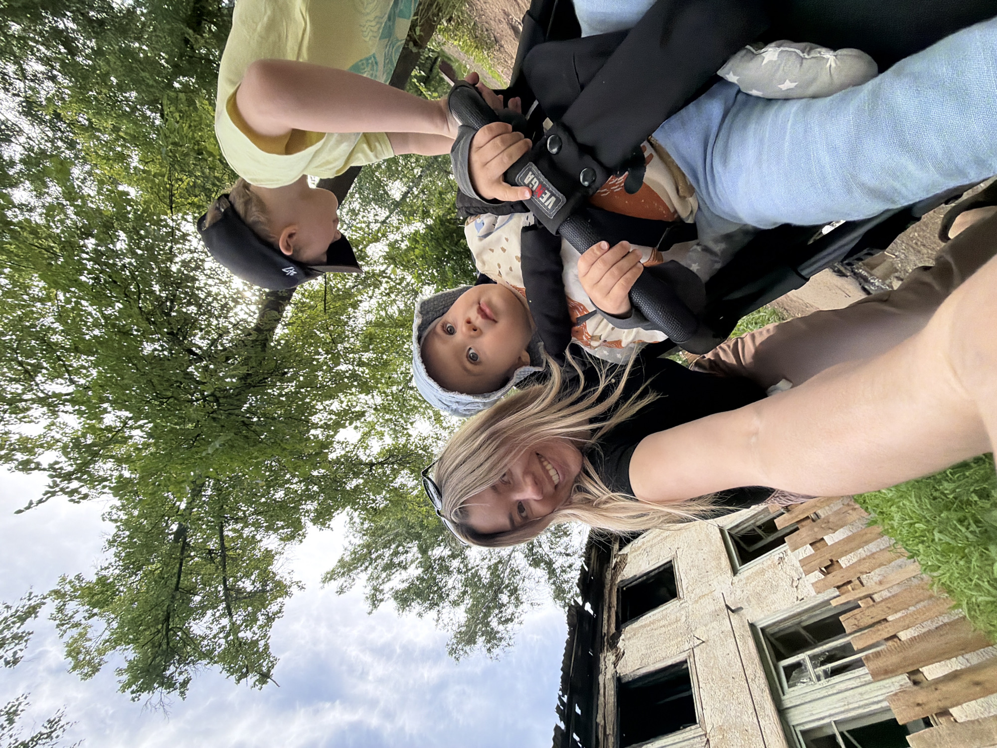 The image, captured from a low-angle perspective, features three individuals in an outdoor setting. On the lower right, a woman with long blonde hair and sunglasses on her head smiles directly at the camera. She wears a dark top, and part of her arm or leg extends into the frame.
To her left, an older child, wearing a yellow t-shirt and a black baseball cap, appears to be looking down or interacting with a baby. The baby, centrally located, has light hair, wears a light-colored hood or hat and a patterned shirt, and is seated in a black stroller. The baby looks directly at the camera with wide, curious eyes. A grey pillow with white stars and a light blue fabric are visible inside the stroller, and the brand name "VEGA" is discernible on a black strap.
The background consists of vibrant green trees and a partly cloudy sky, visible through the foliage, occupying the upper and left sections of the image. On the far right, a damaged building is partially visible, featuring broken or boarded-up windows with wooden planks covering some openings. A patch of green grass is seen adjacent to the distressed building.
The scene depicts a daytime outing, characterized by the happy expressions of the individuals against a backdrop that includes both natural elements and a noticeably damaged man-made structure. The location context for this image is Chaikovsky, Russia.