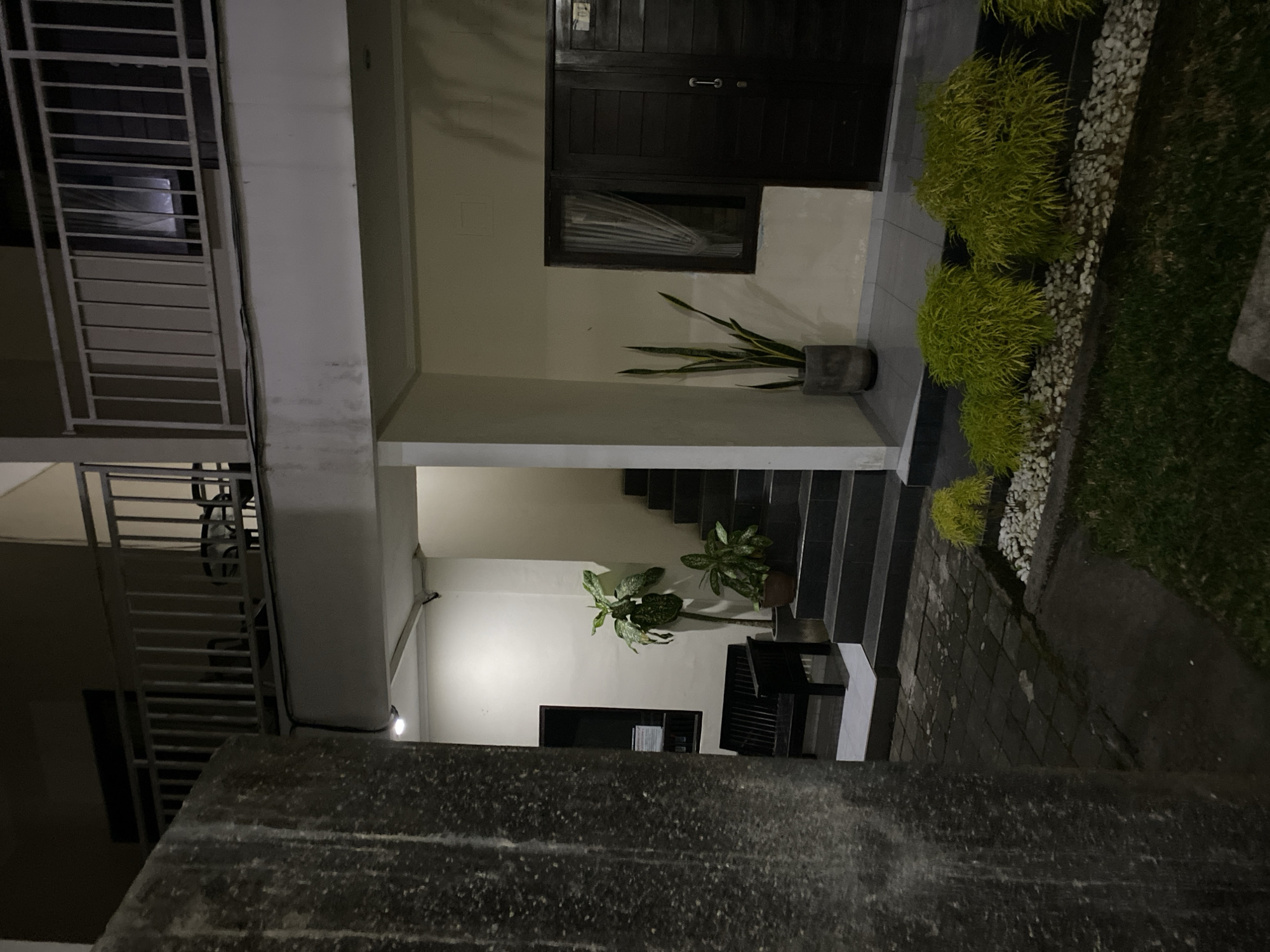 This is an exterior, low-angle, medium shot photograph taken at night in Kuta Selatan, Indonesia. The scene depicts a building with a balcony on the second floor. On the ground level, there is a dark wooden door with a metal handle and a keyhole, and next to it, a dark wooden double door with a small plaque that reads "02". In front of the building, there is a short set of stairs leading up to the doors, with a window to the left of the stairs. A potted snake plant stands to the left of the stairs. To the left of the building, a dark wooden bench sits beneath a window. The ground in front of the building is paved with dark square tiles. In the foreground, there is a patch of green grass and several decorative bushes planted in white gravel. A rough concrete pillar is visible on the far left of the frame. The lighting appears to be artificial, casting shadows and highlighting certain areas of the scene.