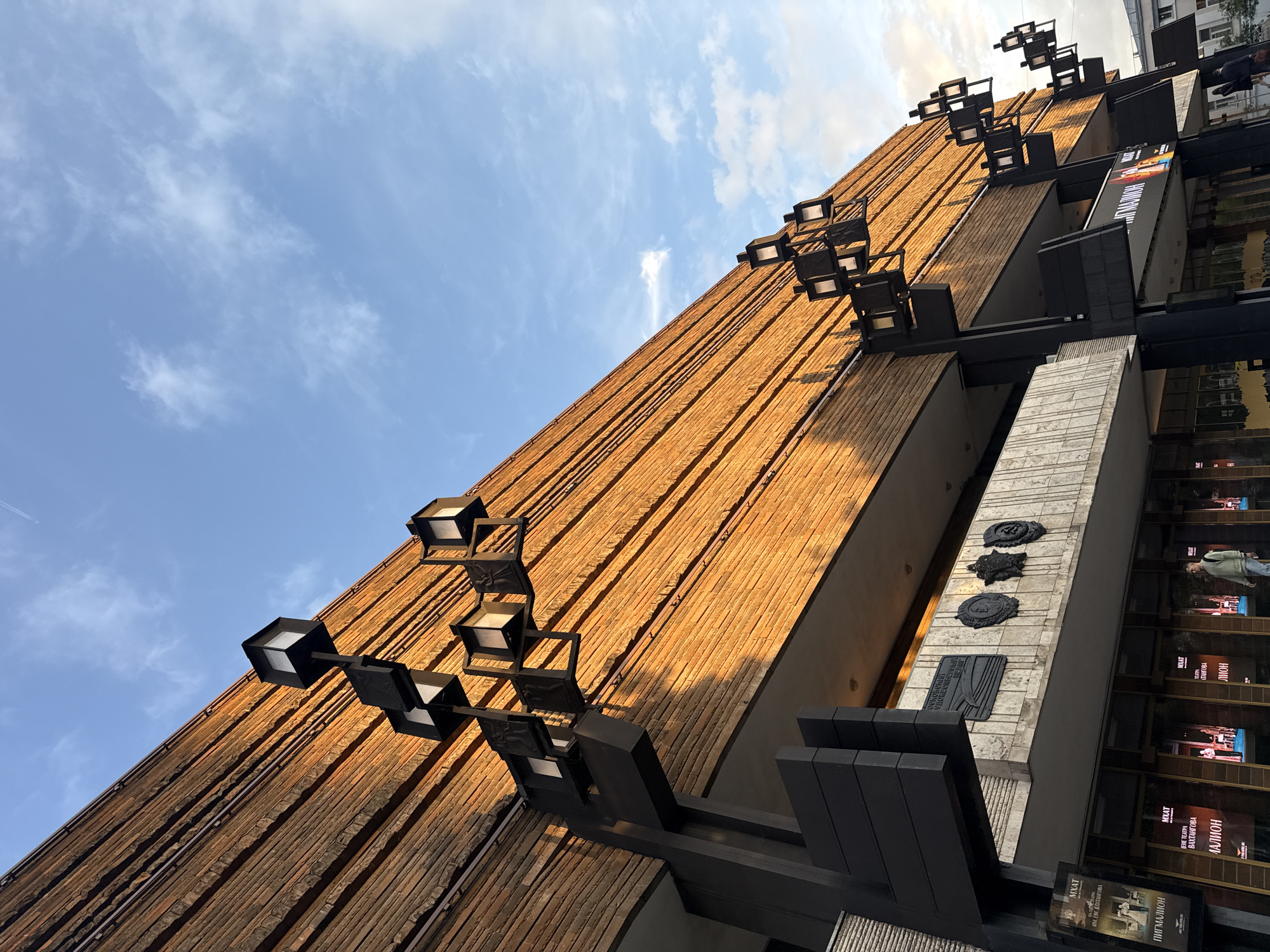 A modern architectural facade rises dramatically against a bright blue sky with scattered wispy clouds, captured from a low-angle perspective. The main section of the building features a warm, reddish-brown textured facade, possibly wood or brick, arranged in strong vertical strips, which are bathed in golden sunlight. Numerous black, cube-shaped light fixtures are mounted on dark metal arms, extending from the facade and casting shadows, creating a geometric pattern.
Below the main textured facade, there are dark, modern structural elements and a lighter-colored, vertically-patterned wall section. On this lighter section, several dark, ornate emblems or plaques are affixed. Below these, large display cases or windows are visible, showcasing theatrical posters. The reflections in these displays faintly show parts of other buildings and abstract human figures.
Visible text on the posters includes "MXAT," which refers to the Moscow Art Theatre, along with Russian words such as "ПИГМАЛИОН" (Pygmalion), "ТЕАТРА" (Theatre), and "СЦЕНА" (Stage), confirming the building's function as a theatrical venue. Other discernible Russian text includes "ЛИОН" (Lyon) and "ИЛЬЯ ДЕРКАЧЁВ" (Ilya Derkachev).
The time of day appears to be daytime, possibly late afternoon, indicated by the vibrant sunlight and distinct shadows. The weather is clear and sunny. A very faint airplane contrail can be seen in the sky in one of the cropped views. The overall scene depicts a section of the Moscow Art Theatre in Moscow, Russia, highlighting its distinctive modern architecture and cultural purpose.