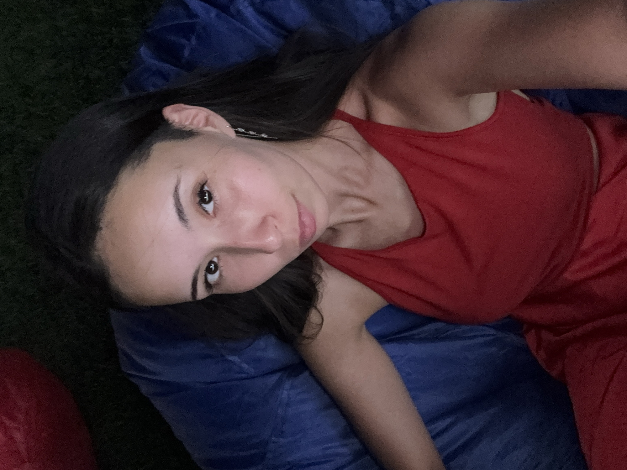 A woman with long, dark hair and brown eyes is lying down, looking up at the camera. She is wearing a red tank top and a silver earring in her left ear. She is on a blue beanbag chair, with a hint of a red beanbag in the upper left corner. The background appears to be grass, suggesting an outdoor setting at night. The lighting is dim, creating shadows on her face and body.