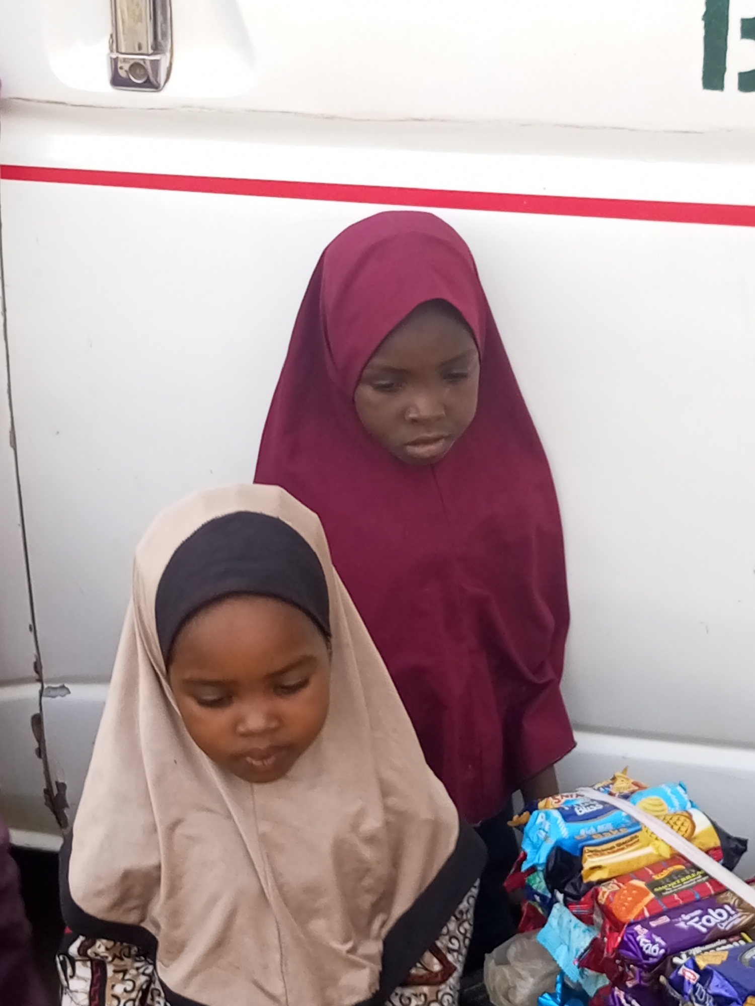 Two young girls wearing hijabs are standing near a pile of packaged snacks. The girl on the left is wearing a beige hijab with a black headband, and the girl on the right is wearing a maroon hijab. The snacks appear to be a variety of biscuits and cookies. The scene is set against a white wall with a red stripe and part of a metal handle. The photograph was taken in Gombe, Nigeria.
