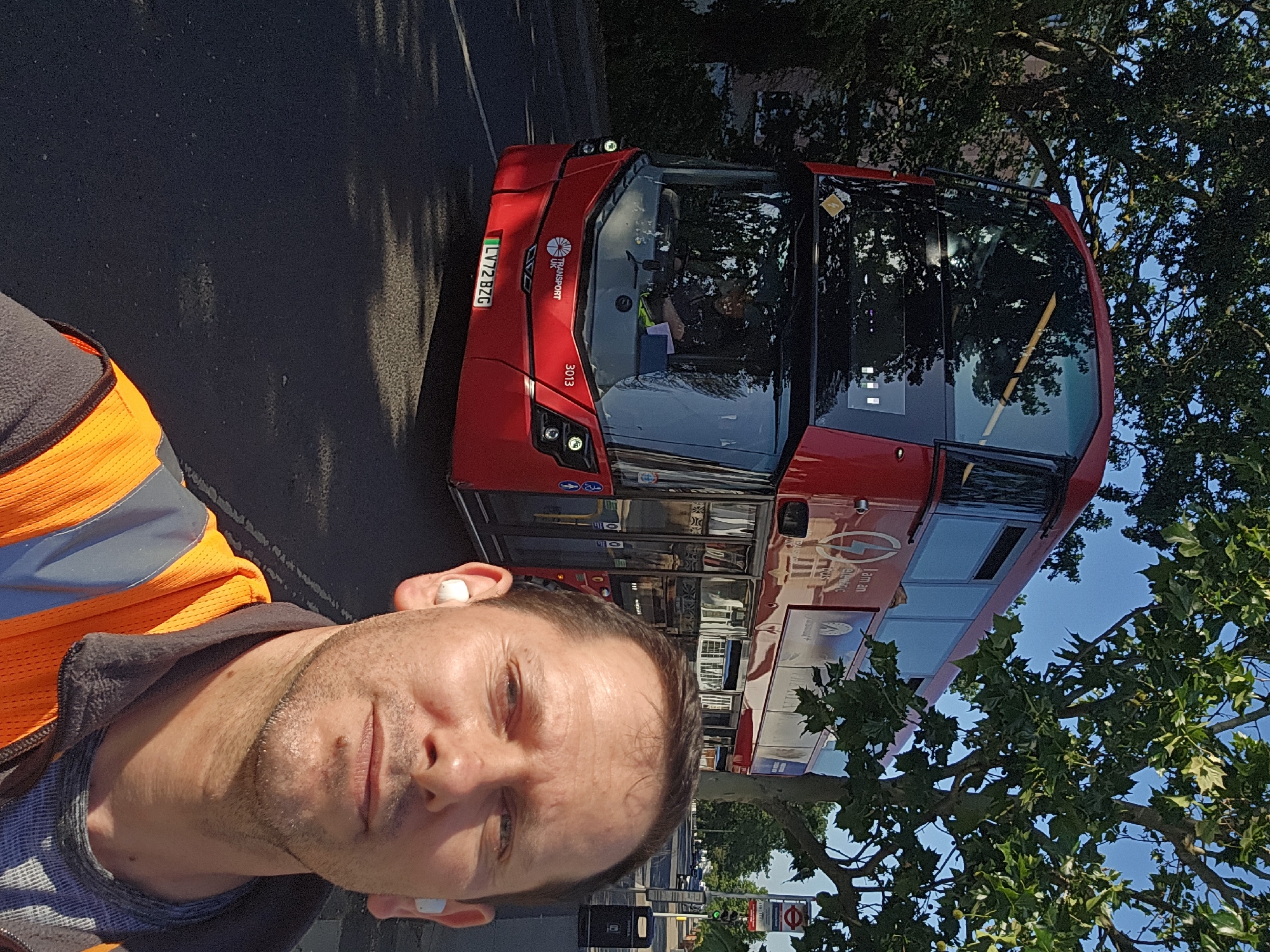 A medium shot of a man wearing an orange high-visibility vest taking a selfie. He has short brown hair and is wearing white earbuds. Behind him is a red double-decker bus with the license plate "LV72 BZG" and the number "3013". The bus is labelled "UK TRANSPORT" with a logo. Trees are visible on the right side of the image, and the sky is blue. A London Underground sign is visible in the background, suggesting the location is London, United Kingdom.