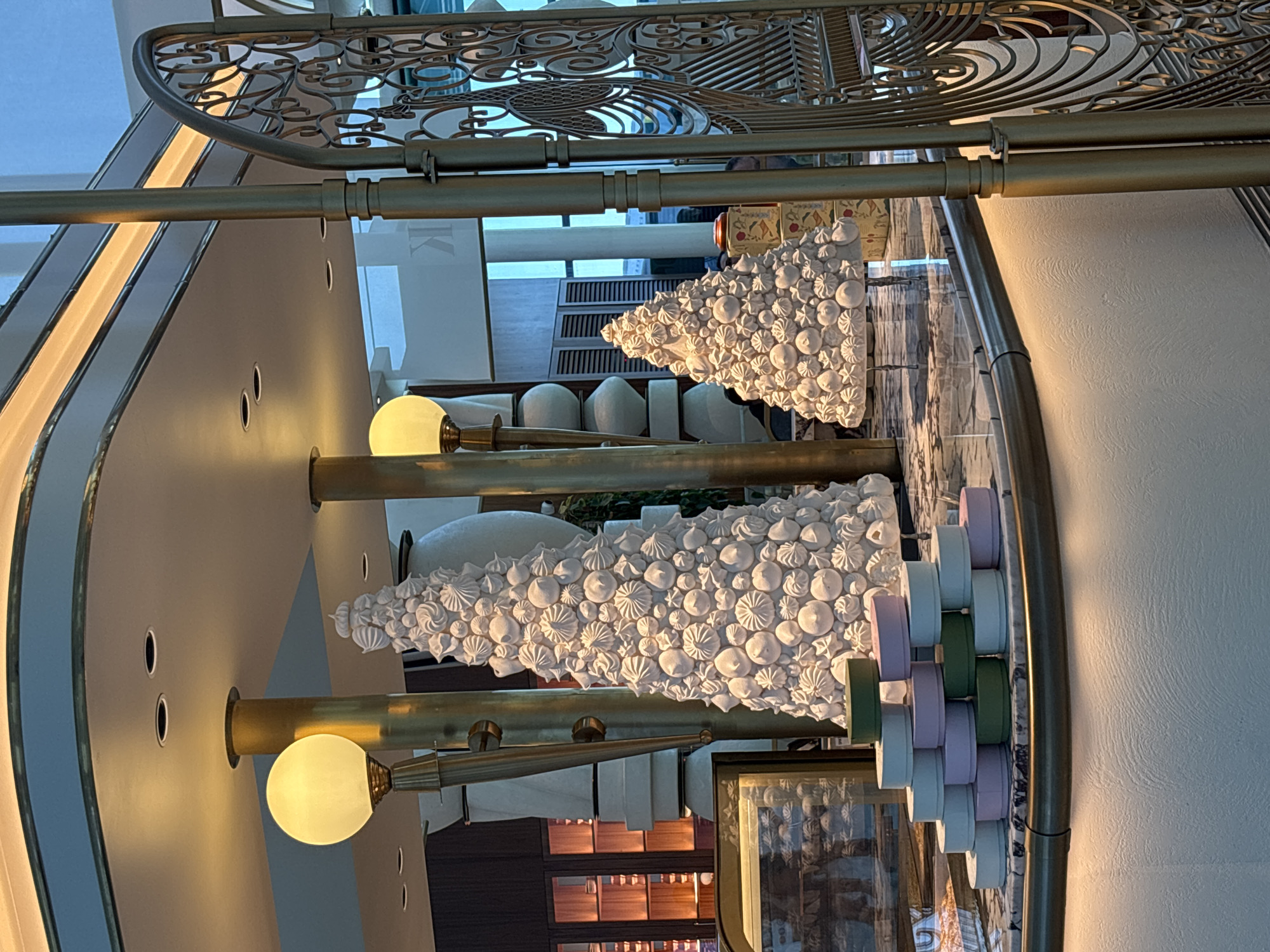 The image captures an indoor scene with two elaborate white meringue Christmas trees, suggesting a festive setting. The larger tree is positioned centrally, while a smaller one stands to its right. In the foreground, a stack of colorful, circular boxes (appearing in shades of green, lilac, and pale blue) is arranged in a pyramid on a marble-topped counter. The counter has a brass trim and is illuminated from below by warm lighting.
Behind the meringue trees, there are tall, golden columns and decorative light fixtures. The background reveals a sophisticated interior with built-in shelving units displaying various items, and large windows with a blueish tint. A decorative golden metal grate or screen is visible on the far right. The floor appears to be polished marble, reflecting the lighting and decor. No people are clearly visible, though a faint figure might be present in the reflection or background. The overall impression is of an upscale establishment, possibly a patisserie, hotel lobby, or a department store during the holiday season, situated in Arnavutköy, Turkey, a district known for its historic waterfront mansions and upscale atmosphere.