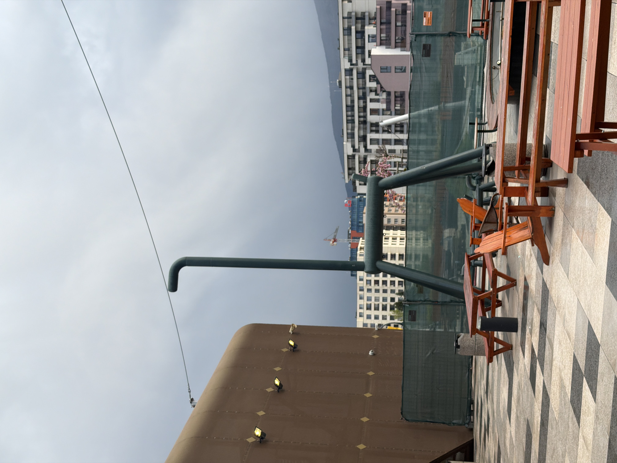 This image captures an outdoor elevated area, likely a rooftop terrace or balcony, overlooking a cityscape under an overcast sky. The foreground features a patterned floor made of black, white, and grey square tiles, and several wooden outdoor tables and benches with reddish-brown staining. A dark, possibly metal, pipe system with an elbow joint extends upwards from the lower left towards the upper right of the frame. To the left, a large, light brown structure with a curved top is visible, fitted with several yellow spotlights that appear to be illuminated. A thin wire or cable stretches diagonally across the sky in the upper left.
In the midground and background, modern-style multi-story buildings, characteristic of urban development, are visible. Some buildings are white and grey, while others have reddish or pinkish accents. A construction crane can be seen between buildings, suggesting ongoing development. A large, dark green mesh fence or netting, possibly for construction or safety, runs along the edge of the terrace. In the distance, a mountain range is discernible beneath the grey, cloudy sky. No people are present, lending a quiet and somewhat desolate atmosphere to the scene. There is no clearly legible text visible within the frame, though a sign on the green mesh fencing is too indistinct to read. The weather is distinctly overcast, and the illuminated spotlights might suggest a dim day or evening setting, though the overall light appears to be daytime. Given the location context, this is a scene from Sofia, Bulgaria.
