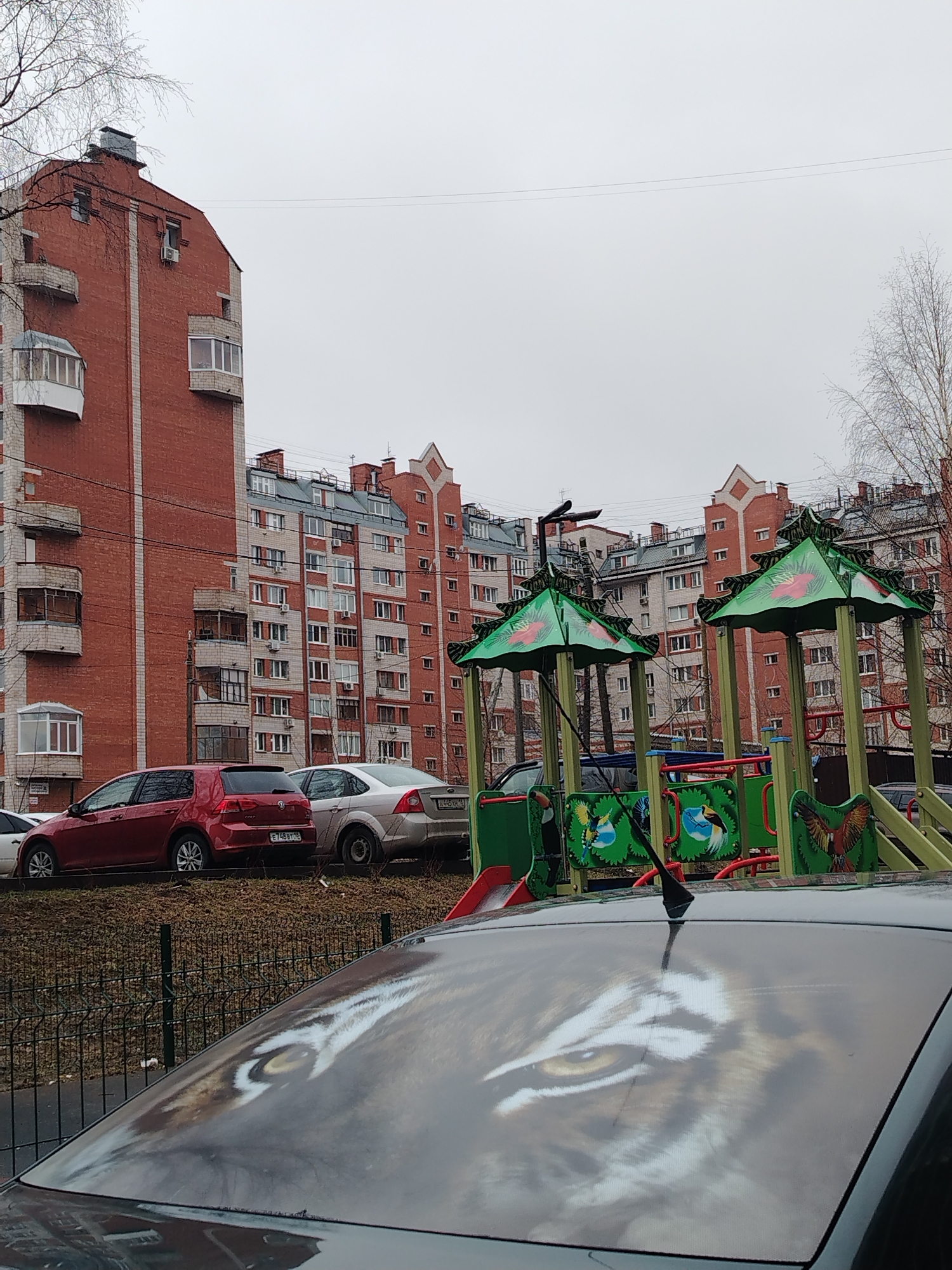 The image depicts a residential area in Izhevsk, Russia, under an overcast sky. In the foreground, the roof of a dark-colored car features a tiger eye design. Behind the car, a small fence separates it from a play area with green-roofed structures decorated with bird motifs. Beyond the play area, a row of parked cars, including a red Volkswagen and a silver Ford, are visible. Further back, multiple multi-story brick apartment buildings dominate the skyline, constructed in the typical Soviet-era style. The season appears to be either late fall or early spring, indicated by the leafless trees.