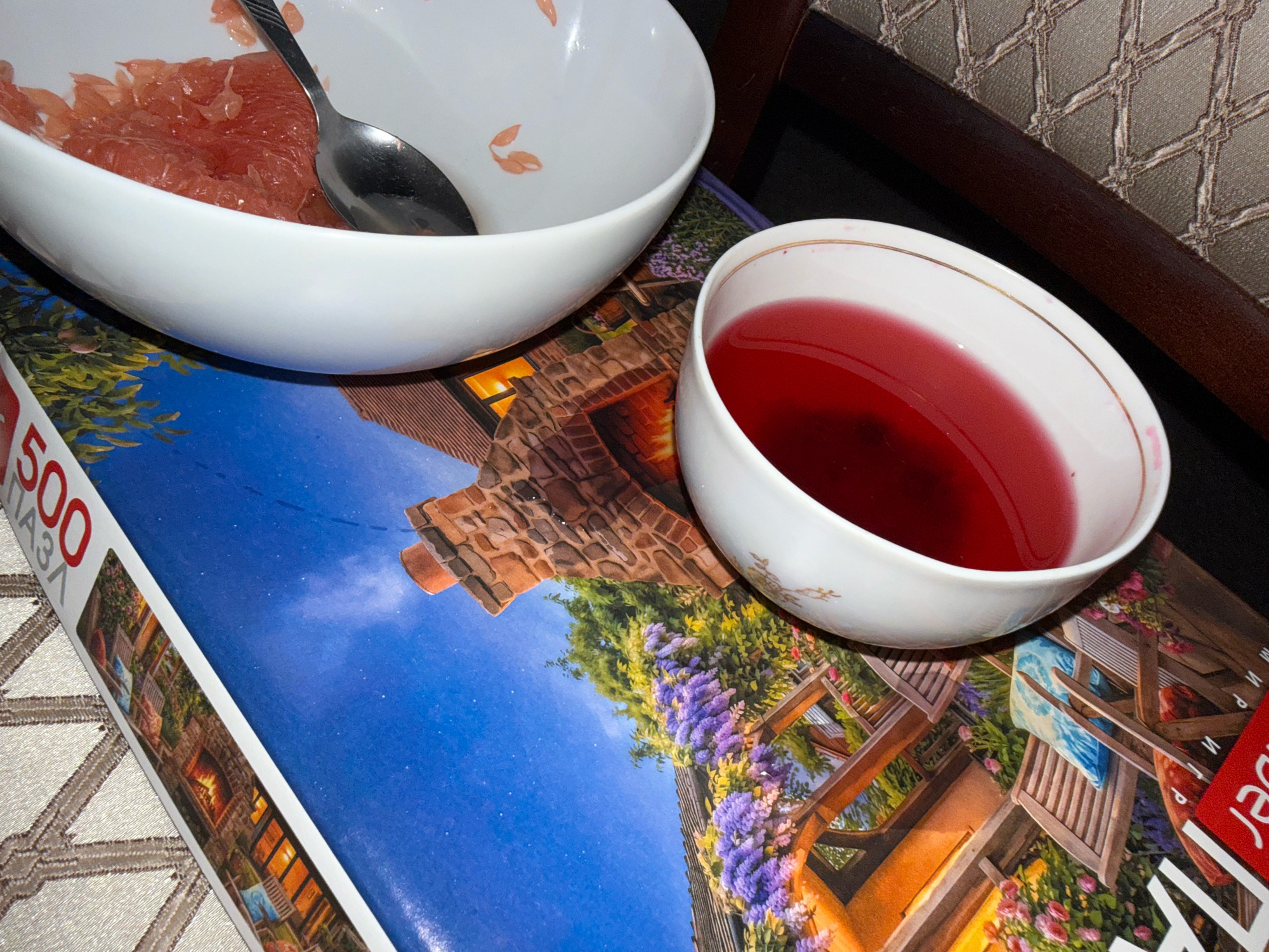 A close-up overhead shot shows two white bowls on top of a puzzle. The bowl on the left contains grapefruit segments and a spoon. The bowl on the right contains a red liquid, possibly tea or juice. The puzzle depicts an outdoor scene with a fireplace and chairs surrounded by flowers. The scene is indoors, with a wooden chair and a textured wall visible in the background. The overall lighting is dim. There are no people visible in the image. The location is not discernible from the image.