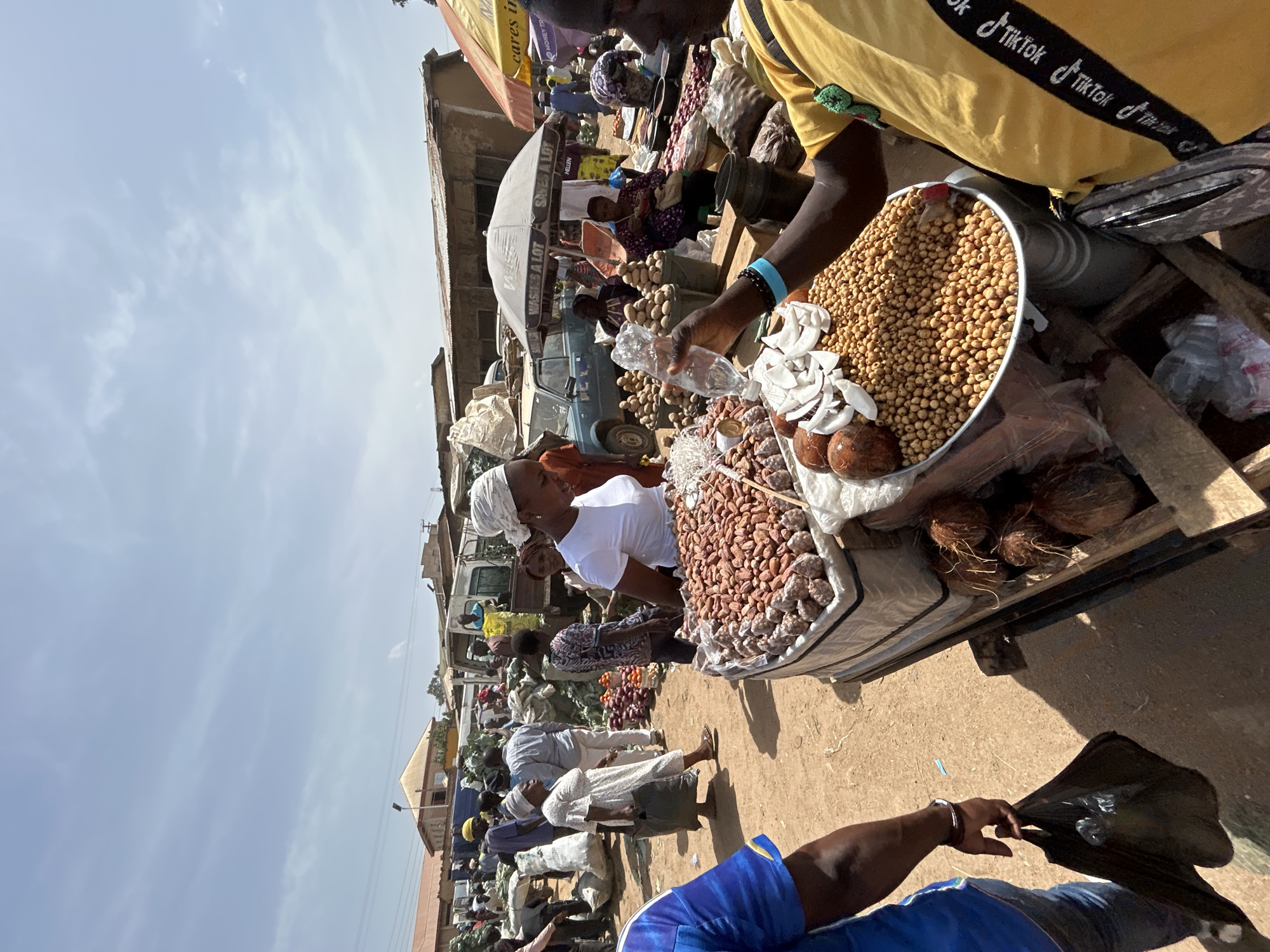 The image depicts a bustling market scene in Bukuru, Nigeria. Vendors are selling peanuts, coconuts, and dates from containers atop a wooden cart. A man wearing a yellow shirt with "Tik Tok" printed on the straps is holding a plastic water bottle over the peanuts. Other vendors and customers, some in traditional clothing, populate the background. The market includes umbrellas for shade and older vehicles. Buildings line the street, with the sky visible above.