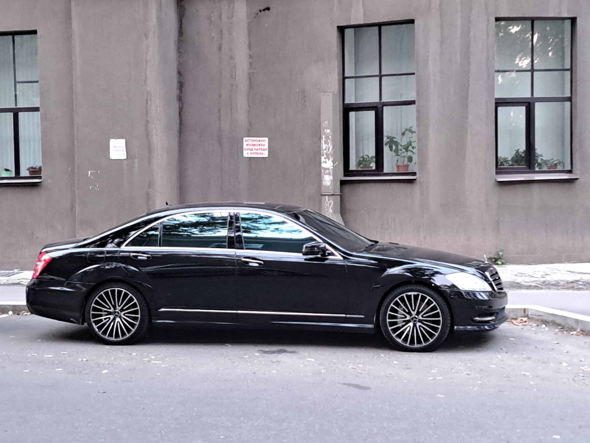 A black Mercedes-Benz S-Class sedan is parked on a paved street next to a gray building. The building has several large windows, some with potted plants visible inside. A small red and white sign is affixed to the building's wall. The street is mostly empty, with some fallen leaves scattered on the asphalt and curb. The image is taken from a slightly elevated side profile, showing the full length of the car. The environment appears to be urban, likely in Irkutsk, Russia, given the architectural style and the presence of a warning sign in Russian. There are no people visible in the image.