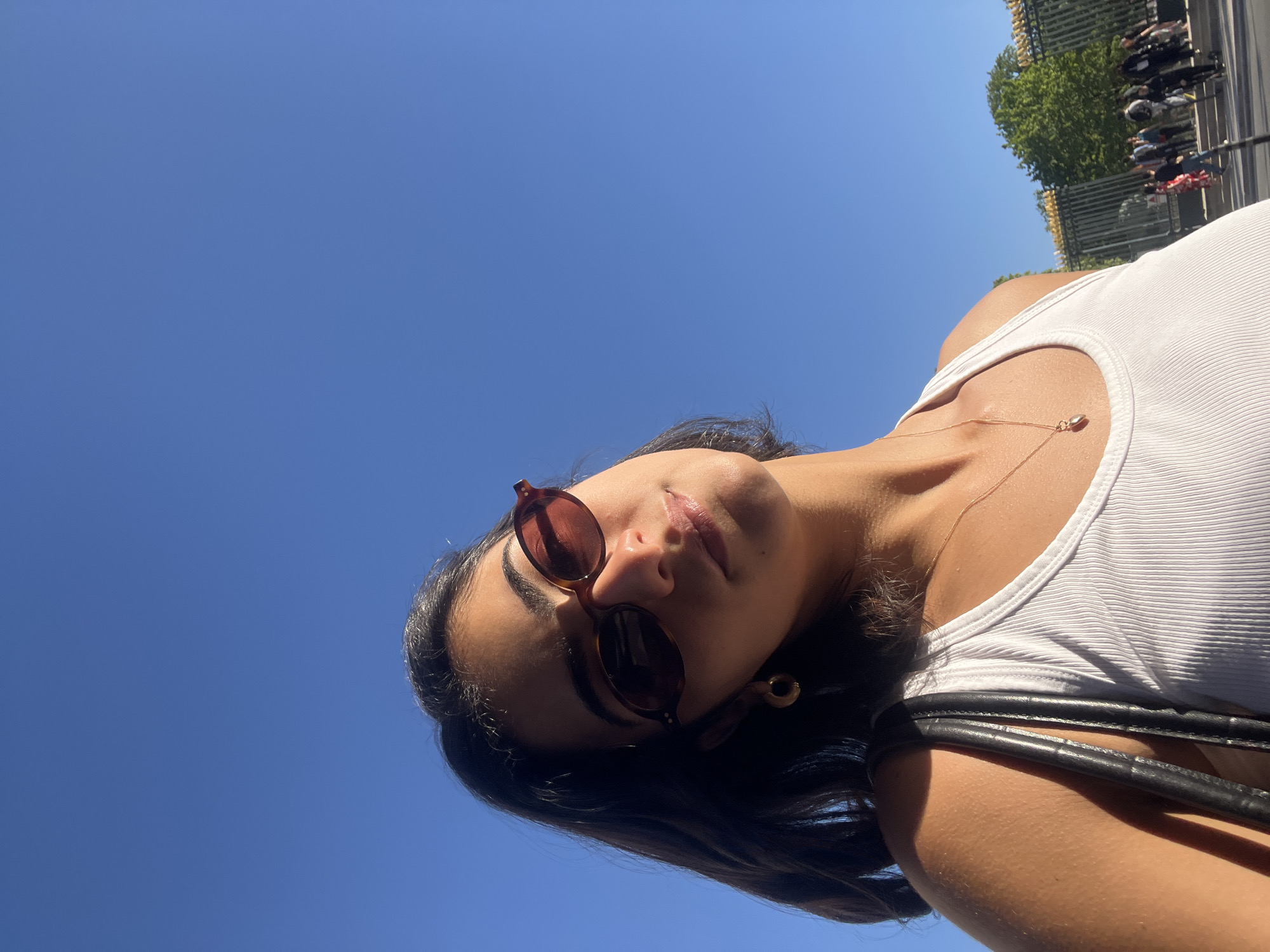 A low-angle, tilted close-up captures a young woman outdoors on a bright, sunny day, likely in an urban setting like Paris, France. She has dark hair and is wearing brown-framed, round sunglasses and a white ribbed tank top. A delicate gold chain necklace with a small pendant adorns her neck, and a black strap, possibly from a bag, is visible over her shoulder. Her tanned skin glows in the sunlight, and her lips are slightly parted, conveying a relaxed and stylish expression as she looks towards the camera. The background is dominated by a clear, vibrant blue sky. In the upper right, blurred elements including green trees, a decorative fence, and distant people suggest a lively outdoor public space. Strong sunlight creates clear shadows and highlights, indicating midday conditions. There is no visible text in the image.