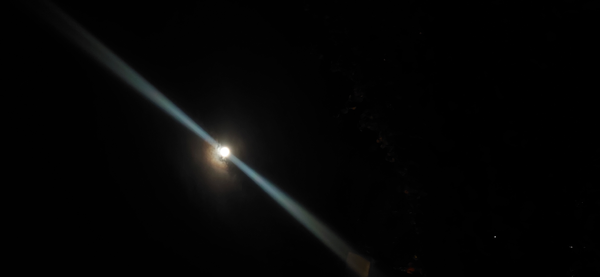 The image captures a night scene with a prominent beam of light cutting diagonally across the frame from the upper left to the lower right. At the center of the beam, a luminous full moon is visible, partially obscured by clouds. The background is predominantly dark, suggesting a nighttime setting. In the lower portion of the image, a faint silhouette of foliage or trees can be discerned against the darkness. The presence of a powerful light source and the moon suggests an urban or semi-urban environment where artificial light might interact with natural celestial bodies. The overall impression is of a dramatic nocturnal illumination. The location of Presidente Franco, Paraguay, is not directly evident from the visual cues alone but would require contextual information to be associated with the scene.