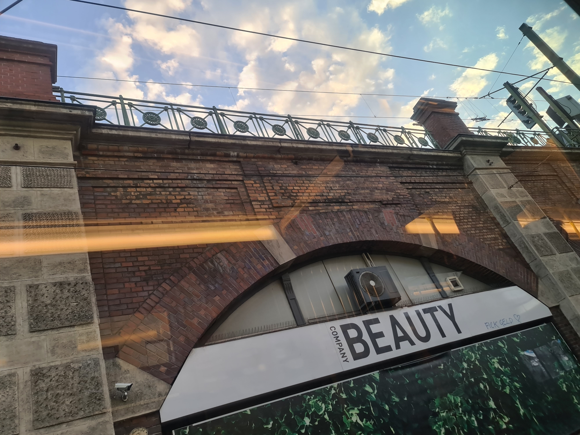 The image shows a close-up view of a brick archway under a bridge or elevated structure. The sky above is blue with scattered white clouds, and there are power lines visible. The brickwork is aged and shows some signs of wear.
Below the archway, a white sign with black text reads "COMPANY BEAUTY". To the right of this, handwritten in blue, is "FICK GELD ♡". There is also an air conditioning unit mounted on the wall above the sign.
On the left side of the archway, there is a grey stone wall with textured blocks and a security camera mounted on it. The overall impression is an urban setting, likely within a city, with a slightly gritty, industrial feel. The lighting suggests it is daytime, possibly late afternoon or early evening due to the warm tones in the sky.
While the exact location in Vienna, Austria is not explicitly stated, the graffiti "FICK GELD" (German for "fuck money") is a common expression that could be found in various urban environments. The presence of what appears to be a tram or train line overhead, indicated by the power lines and overhead equipment, suggests a transportation hub or route within the city.
