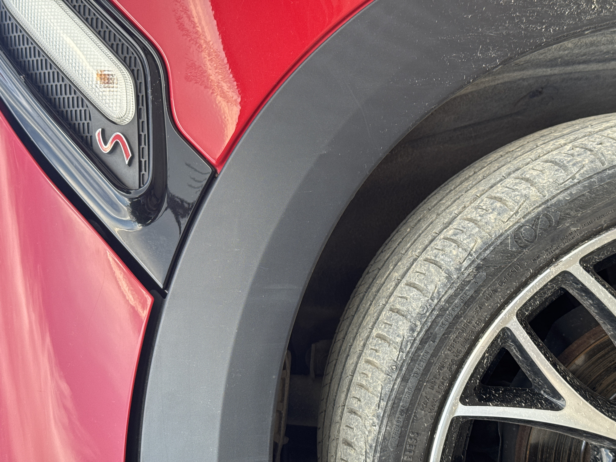 This media file is a close-up, eye-level shot of a red car's wheel well, focusing on the fender, tire, and part of the car's body. The car is red and has a black plastic fender flare. A red "S" emblem is visible on the black trim next to a turn signal light. The tire is dirty, indicating it has been driven on dusty roads. The rim of the wheel is black and silver with a multi-spoke design. The scene appears to be outdoors during the day, with natural light illuminating the car. The background is blurred, but it suggests an outdoor setting, possibly a road or parking area. There is no visible text in the immediate foreground, but some tire information is visible on the sidewall of the tire. The overall impression is a detailed, focused view of a part of a car, emphasizing its design and condition. The provided location context of Radanovići, Montenegro, suggests this is where the image was taken.