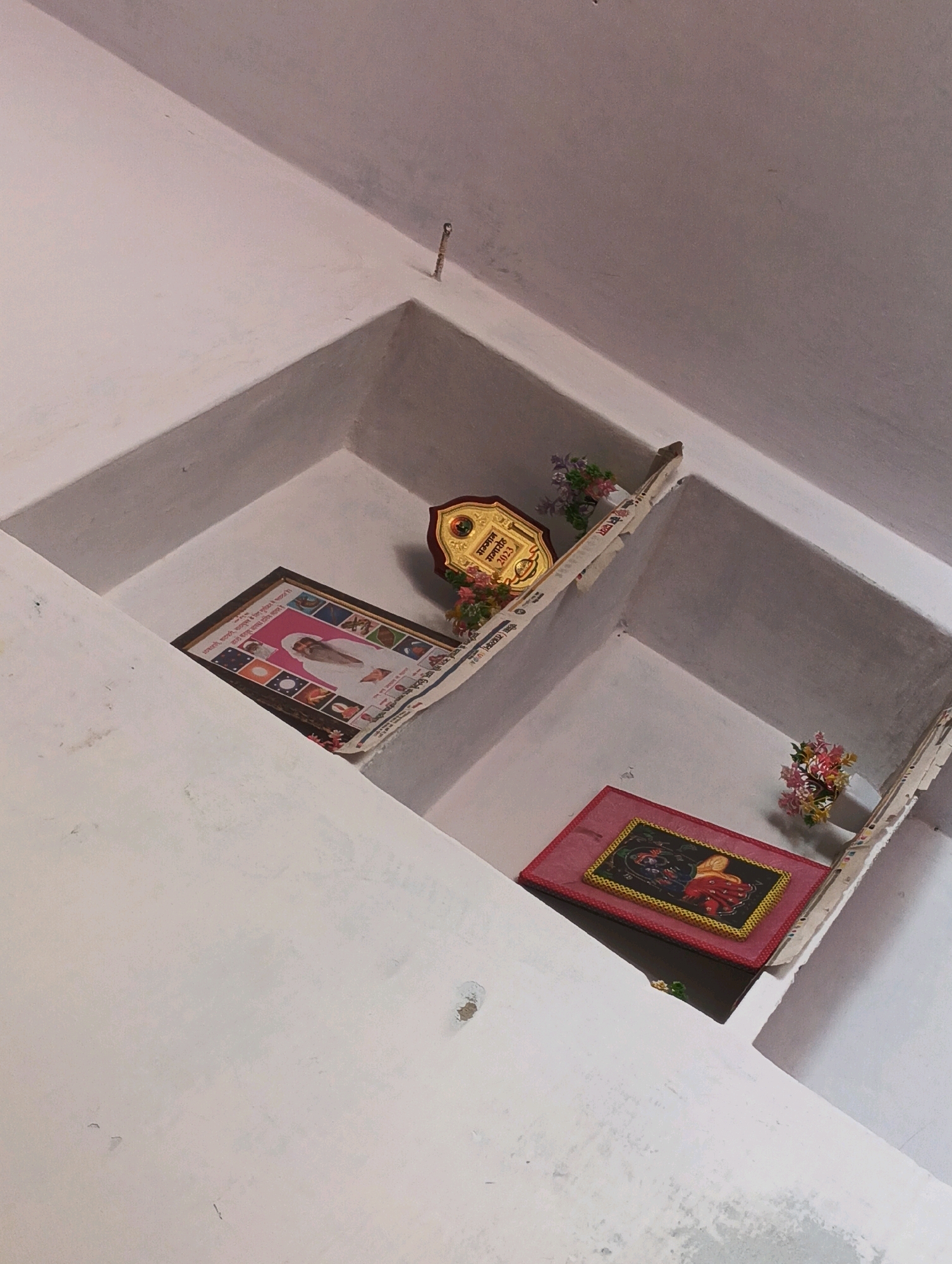 The image shows a white, built-in shelf unit in a room. On the top shelf, there is a gold plaque with text and some artificial flowers. To the left of the plaque is a framed picture of a person in white robes, surrounded by smaller images. This picture is placed on top of some folded newspaper. On the lower shelf, there is a red framed object with a decorative image of a figure in red and yellow. There are also some artificial flowers placed on the edge of this shelf. The walls of the shelf unit appear to be made of plaster and are painted white. The overall environment suggests a storage or decorative space. The city of Kurli, India is not visible in this image, nor are there any people present.