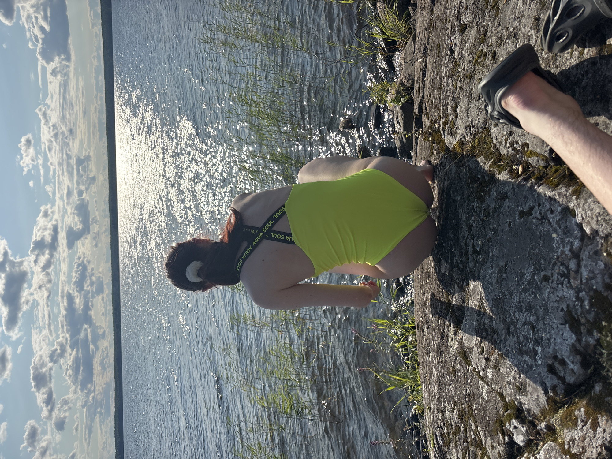 A person wearing a neon yellow one-piece swimsuit is squatting on a rocky shoreline, facing away from the camera and towards a large body of water. The water is sparkling with sunlight reflecting off its surface, and a tree line is visible in the distance. Tall reeds grow along the edge of the water. In the foreground, a person's foot wearing a black Croc is visible, resting on the rocks. The sky is blue with scattered white clouds. The scene appears to be a natural outdoor setting, likely a lake or a large river. This location is within the city of Shuiskaya Chupa, Russia.