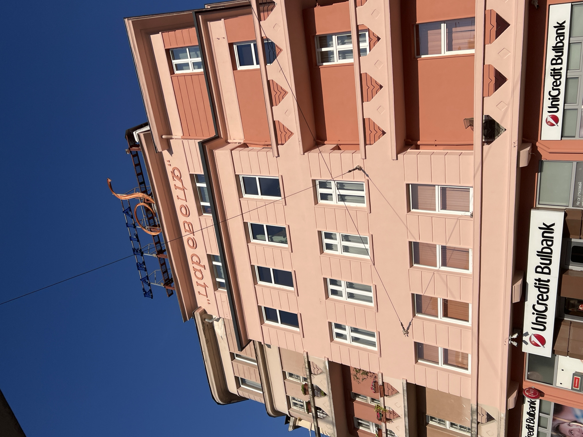 The image captures a low-angle view of the facade of a building in Sofia, Bulgaria. The building exhibits a distinctive pink and coral color scheme, with multiple floors featuring numerous windows. Several of the windows are adorned with white frames and sheer curtains, while others are dark, reflecting the sky. The upper portion of the building displays a prominent sign with blue lettering that reads "KARIBIA" and another sign in Cyrillic script that appears to say "ЦАРЕВЕЦ". A decorative, orange, curved metal sculpture is also visible on the roofline. The lower section of the building features storefronts, most notably displaying the "UniCredit Bulbank" logo. The sky above is a clear, vibrant blue. Power lines are strung across the scene, partially obscuring the upper parts of the building. The architecture suggests a commercial or residential building likely situated in a city center.