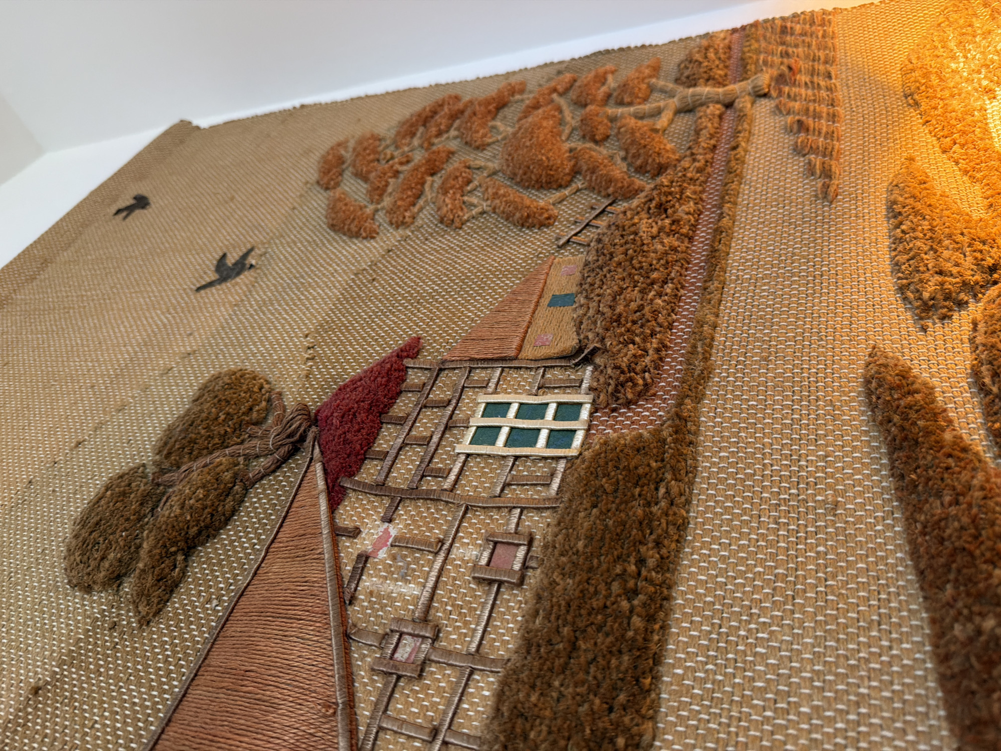 A close-up, angled view of an embroidered tapestry depicts a rural landscape. The central subject is a brown, Tudor-style house with a prominent red, loop-pile roof and a green-paned window, outlined with white threads. The house walls are rendered in intricate brown and white woven patterns with dark brown simulated timber framing. To the left of the house, a cluster of shaggy brown trees or wheat bundles rises. Two dark bird silhouettes are visible in the upper right sky area. To the right of the house, a large, shaggy brown tree with multiple branches extends upwards, rendered in a dense, loop-pile texture. The ground surrounding the house and trees is also depicted with varied textures of brown yarn, suggesting foliage or terrain. The tapestry's base material is a light tan, tightly woven fabric with a visible grid pattern of white horizontal threads over brown vertical threads. Warm, yellow light illuminates the lower right portion of the tapestry. The scene is a stylized, artistic depiction within a textile, not a physical location in a city.