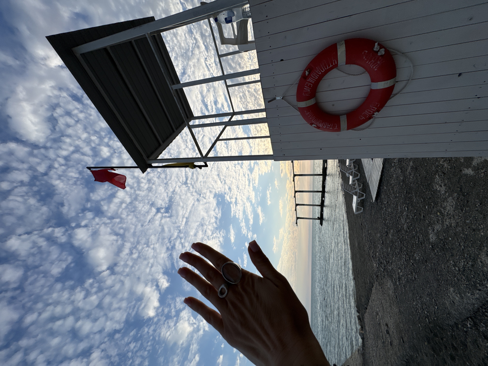 A hand with two rings is raised in the foreground, obscuring part of a beach scene. Behind the hand, a red lifeguard buoy hangs on a white wooden structure, likely a lifeguard station. The station has a metal roof and a red flag is flying from a pole. The sea is visible in the background, with two sun loungers on the beach and a pier in the distance. The sky is filled with scattered white clouds. The scene is likely the city of Sochi, Russia.