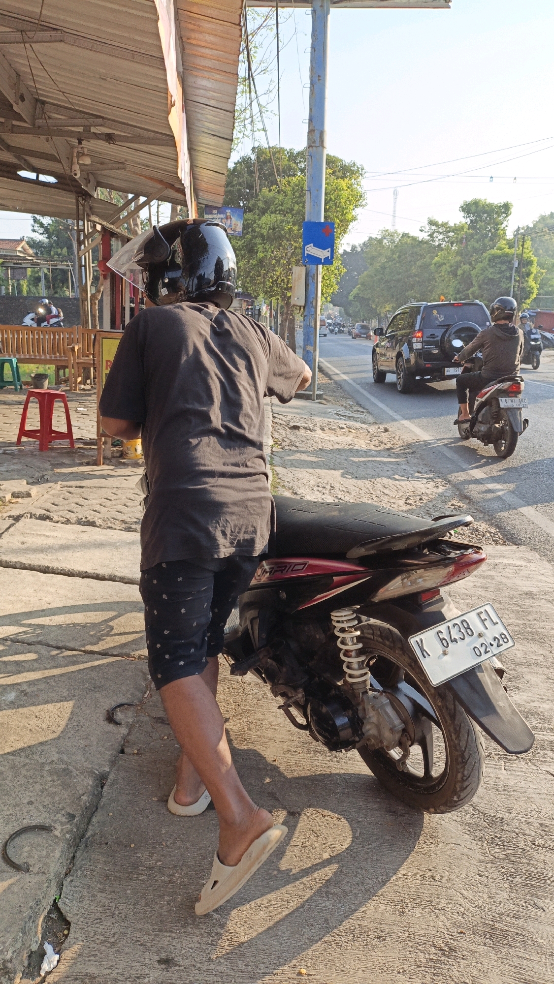 A man wearing a black t-shirt, dark patterned shorts, a black helmet, and light-colored slip-on shoes stands next to a red and black motorcycle on a concrete sidewalk. The motorcycle's license plate reads "K 6438 FL". The man is facing away from the camera and appears to be either getting on or off the motorcycle. In the background, there is a street with several vehicles, including a dark SUV and another motorcycle with a rider. Trees line the street, and the sky is bright with sunlight. To the left of the man, there is a covered seating area with wooden benches, a red stool, and some foliage. A blue sign with a white medical cross symbol is visible on a pole. The overall scene depicts a street-side view in what appears to be a semi-urban or commercial area of Kuwasen, Indonesia.