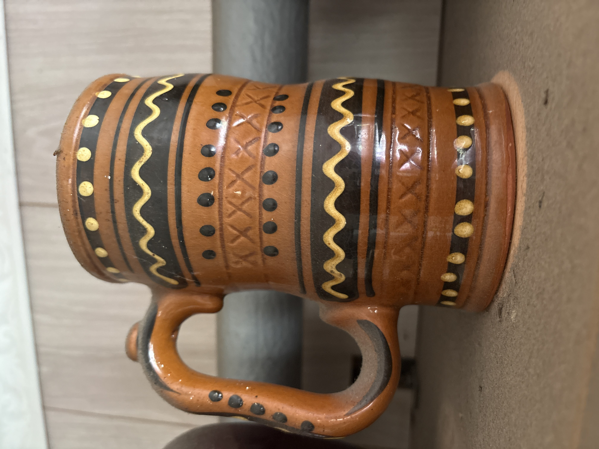 A close-up, eye-level, slightly low-angle shot captures a rustic, earthenware mug with a handle. The mug is decorated with horizontal bands of black and yellow wavy lines, yellow dots, and black dots. A crisscross pattern is incised into the orange-brown surface. The mug appears dusty, suggesting it is in a storage area. The background is out of focus, showing a shelf or surface. The overall impression is of an aged, possibly handmade item. The location of Ветлужский, Russia is not determinable from the visual information provided in the image.
