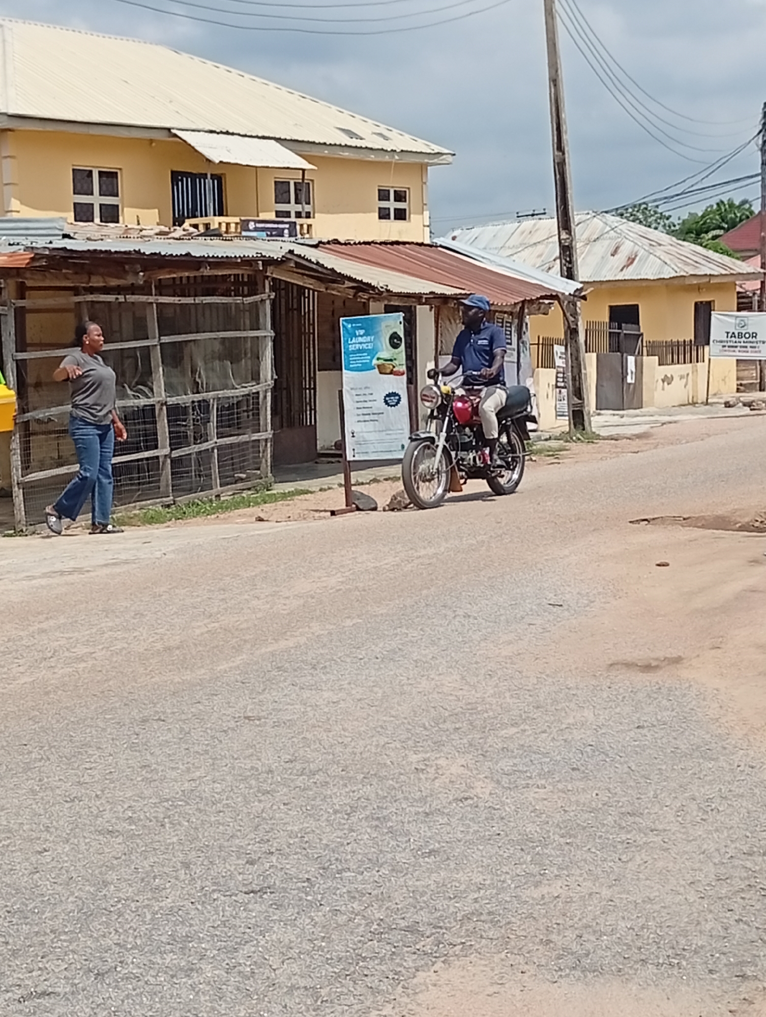 The image shows a street scene in Lokoja, Nigeria. In the foreground, a street stretches into the distance. On the left, a woman is walking on the sidewalk next to a structure with a corrugated metal roof and wire mesh siding. Beside the structure stands a sign advertising "VIP Laundry Service." A man on a red motorcycle is approaching the woman in the middle of the street. In the background, two buildings with light-colored walls and corrugated metal roofs are visible. Wires and a utility pole are visible against a cloudy sky. A sign that reads "Tabor Christian Ministry" is on the side of a building.