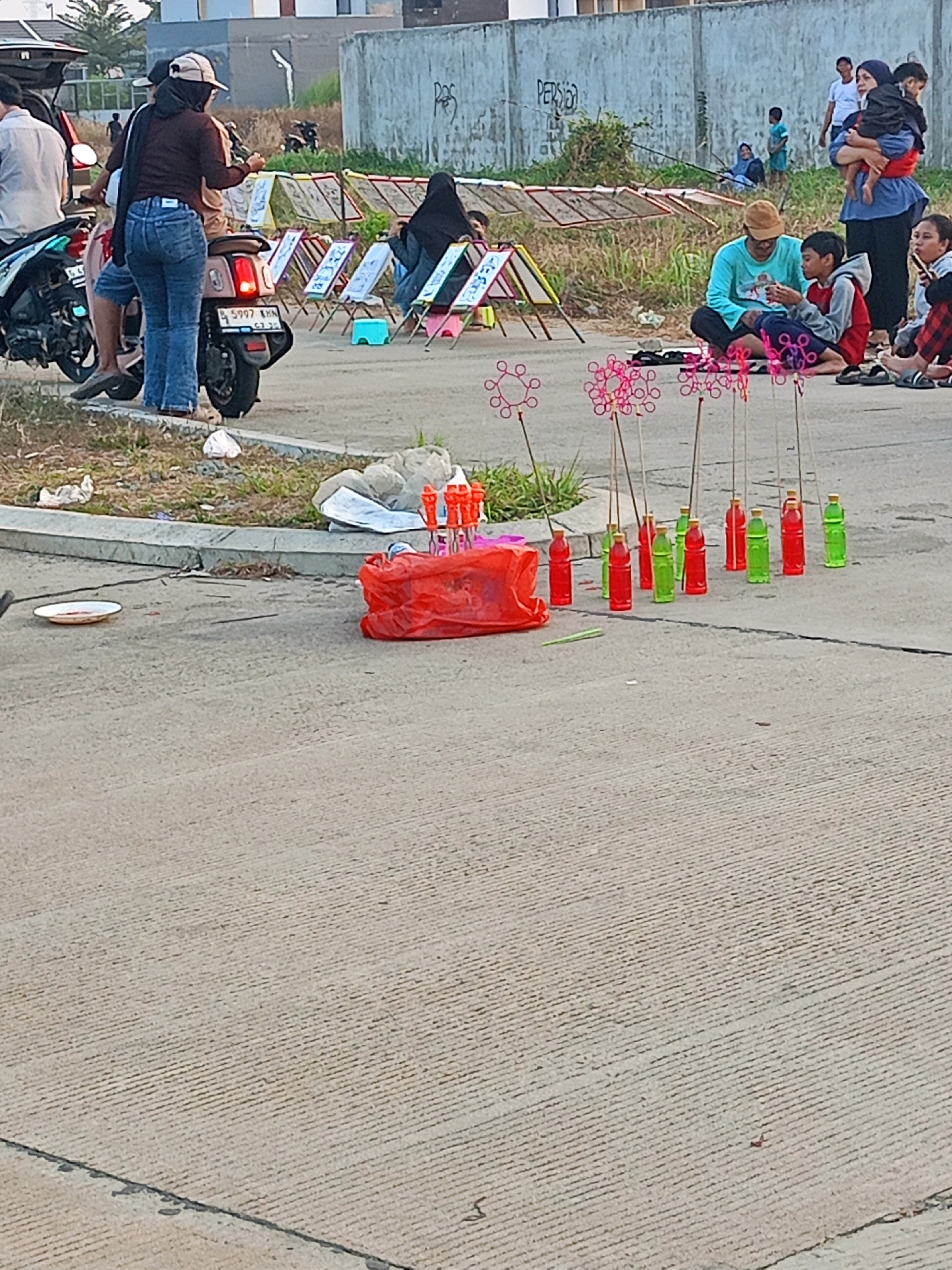 The image depicts an outdoor scene in an urban environment, possibly a street or a roadside area, under daylight. In the foreground, a collection of items are laid out on the pavement, including several red and green plastic bottles, some with yellow caps, standing upright on sticks, suggesting they might be part of a game or a display. In front of these bottles is a large red plastic bag. Further to the left, there is a white plate with some reddish residue on it.
In the middle ground, several individuals are present. A woman wearing blue jeans and a brown top, with a tan cap, is seated on a scooter, with another person behind her. To the right, a group of people are gathered. A man in a light blue shirt is sitting on the ground, interacting with a boy wearing a red and gray shirt. A woman in a blue shirt and headscarf is holding a baby. Other children are also visible in this area. Behind this group, there are several framed displays, possibly for sale or exhibition, and some drying racks with material stretched across them. A black motorcycle is parked to the left.
The background shows a concrete wall with graffiti on it and some vegetation. Other people, including children, are visible further in the distance, some near what appears to be a structure or a fence. The overall setting suggests a casual gathering or a small market-like activity.
