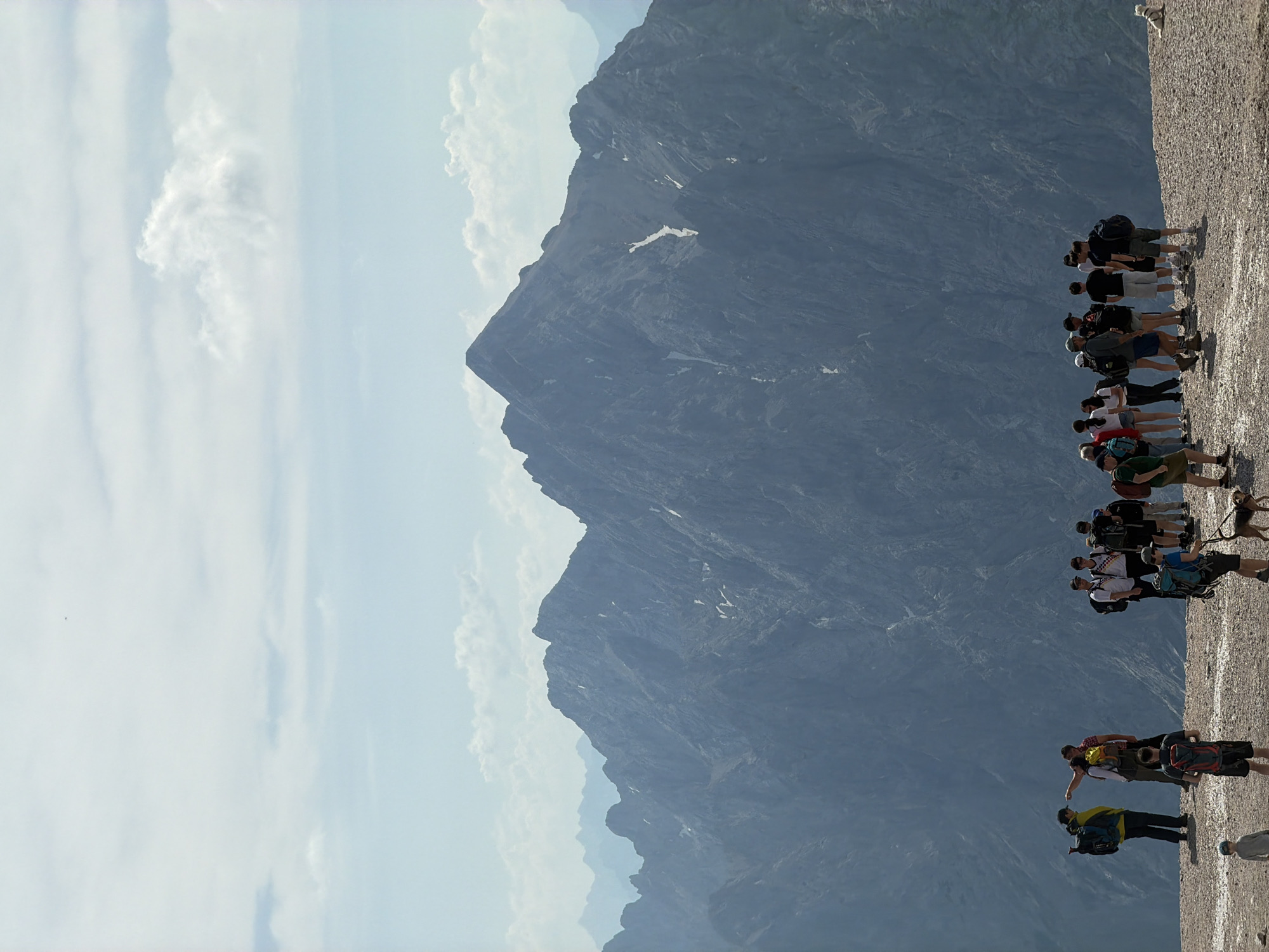 The image depicts a group of hikers at a mountain summit, likely in the Bavarian Alps near Garmisch-Partenkirchen, Germany, given the characteristic jagged peaks and clear skies. The foreground features a gravelly surface where several individuals and a dog are present. A prominent group of about a dozen hikers stands gathered, some with backpacks, appearing to be resting or observing the view. To the left, two hikers are interacting, with one pointing towards the distance. Further to the left and slightly in front, a young boy stands. In the mid-ground, a large, imposing mountain range dominates the scene, its rocky slopes partially covered in snow. The sky above is a pale blue with scattered white clouds. The overall atmosphere suggests a scenic and recreational mountain environment.