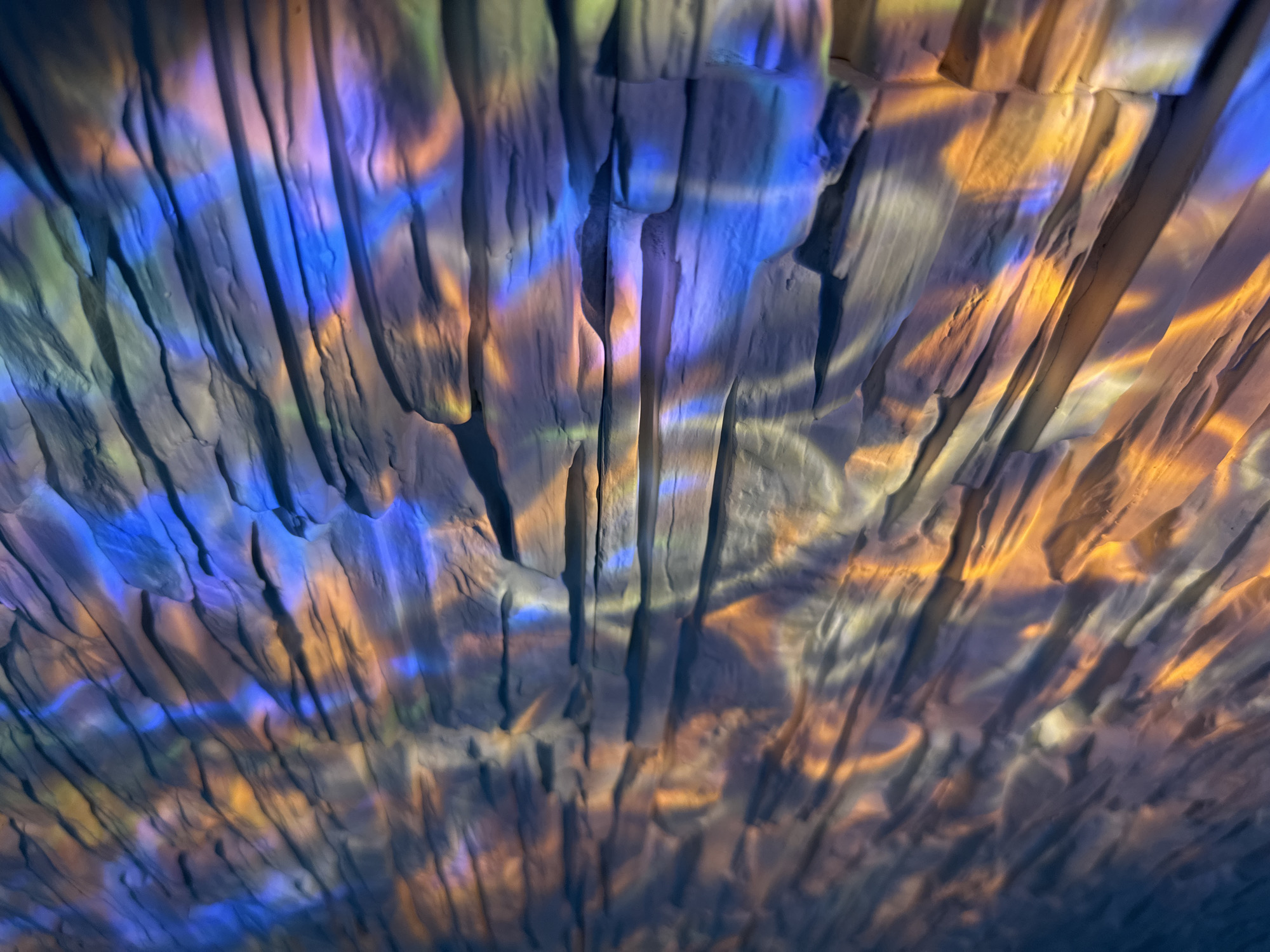 The image depicts a textured surface with a pattern of vertical striations, resembling rough-hewn rock or wood. The surface is illuminated with multicolored light, predominantly blue on the left side and transitioning to orange-yellow on the right. There is a play of shadows and highlights that accentuate the texture. The location is Unknown, Unknown. No people, subjects, or actions are visible in the image.