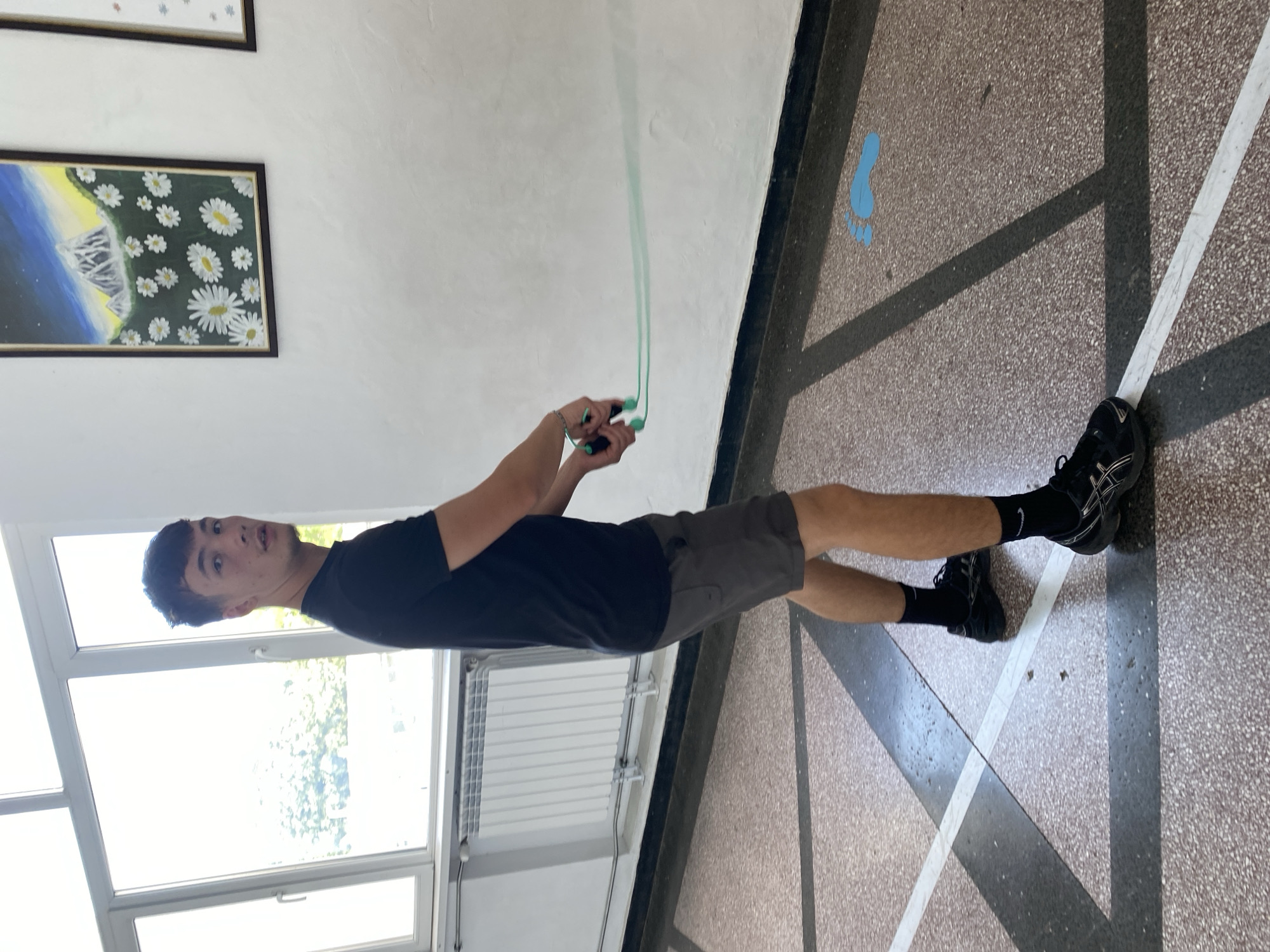 A young man stands in an indoor space holding a jump rope. He is wearing a black t-shirt, grey shorts, black socks, and black running shoes. Behind him are windows and a radiator. On the wall are two framed paintings. The floor is tiled with a geometric pattern, including a painted blue footprint. The location is likely an interior hallway based on the appearance of the tiles, paint, and architectural style. The city and specific building in Varna, Bulgaria cannot be determined from the visual information provided.