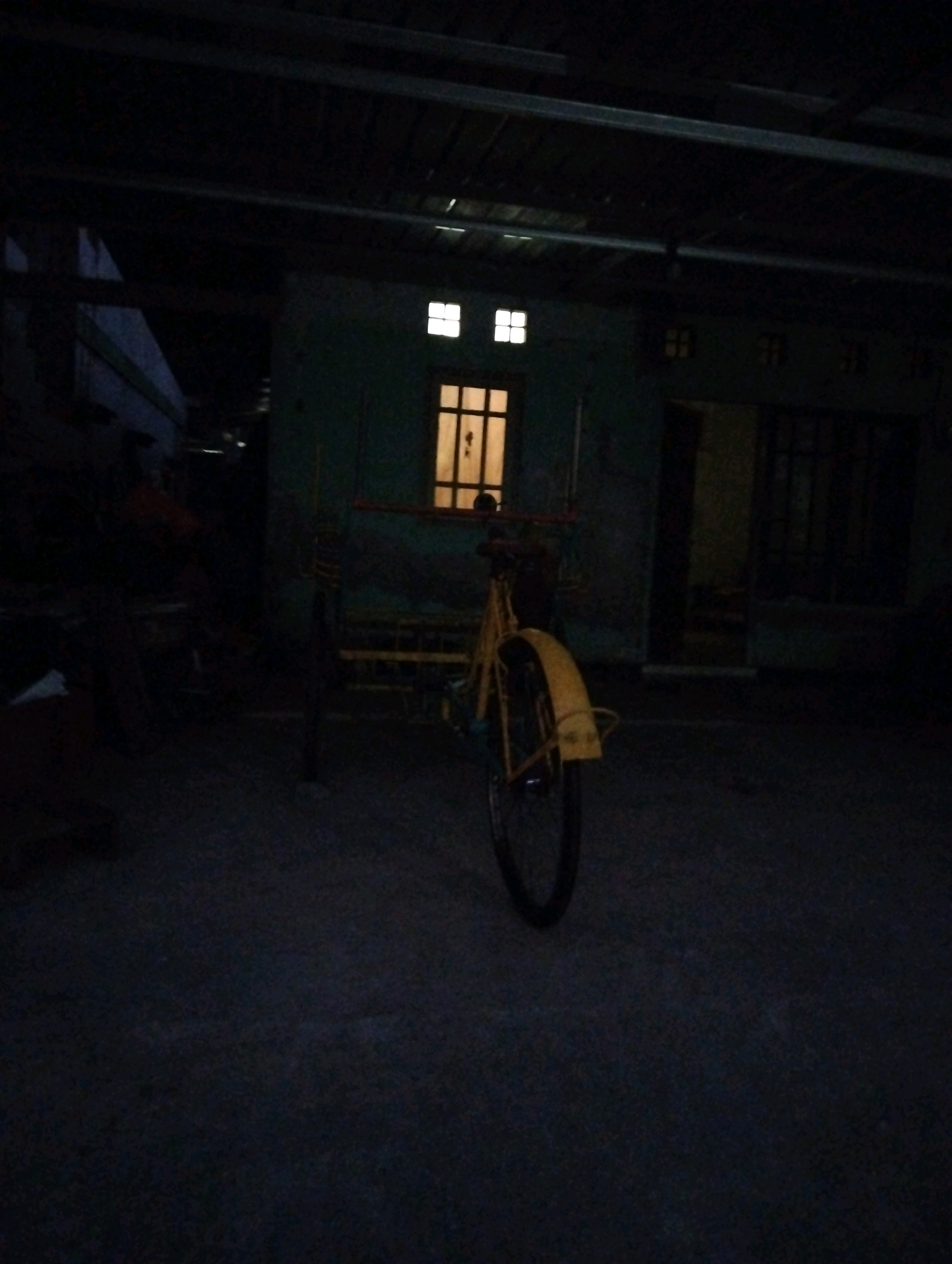 The image depicts a dark, outdoor scene, likely a yard or alleyway, in front of a building with a green wall and windows. A yellow tricycle with a spoked wheel is the central subject, positioned on a paved or concrete surface. The tricycle's frame and rear fender are visible. The building appears to have a corrugated metal roof structure overhead, creating a shadowed, enclosed environment. One window in the building emits a warm, yellow light. The overall impression is one of low light conditions, suggesting it is either nighttime or a dimly lit area. The scene does not provide specific geographic markers to confirm its location in Salatiga, Indonesia, but the architectural style and the presence of the tricycle are consistent with residential areas in Southeast Asia.