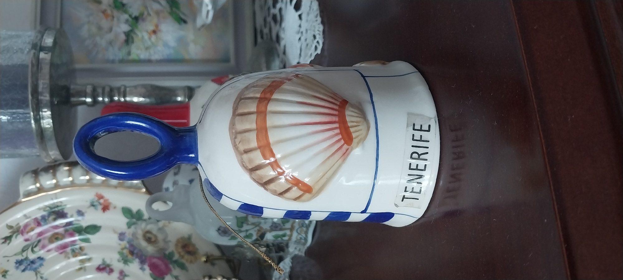 A ceramic bell with a blue handle and a painted seashell on its front sits on a dark brown wooden surface. The bell has blue stripes near its top and a label that reads "TENERIFE". The background is blurred but shows a floral patterned plate, a silver candlestick, and a framed picture. The bell is the primary subject, positioned in the center of the frame. The lighting is soft, and the overall scene appears to be indoors, possibly in a domestic setting. The location in Moscow, Russia is not determinable from the image.