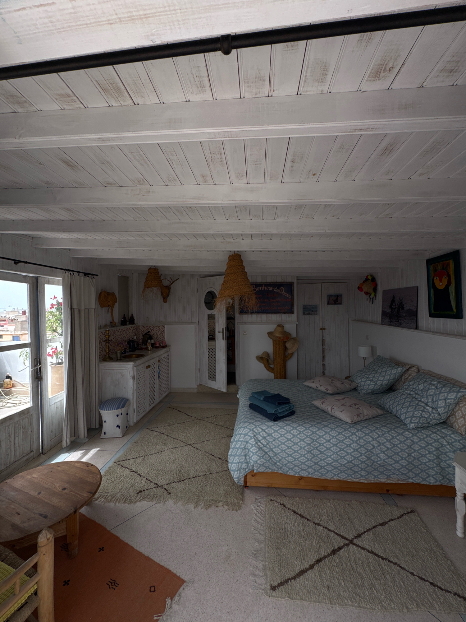 The image depicts the interior of a room in Essaouira, Morocco, likely a guesthouse or rental property, featuring a blend of bohemian and coastal design elements. The room is characterized by its bright, airy feel, with whitewashed wooden paneling on the ceiling and walls.
In the foreground, a rustic wooden dining table with a light-colored base is positioned on the left side of the frame. Adjacent to it, a section of a wicker chair with a yellow cushion is visible. The floor is covered with a light-colored, textured rug with a geometric dark brown pattern, overlapping with a smaller rust-colored rug adorned with dark brown symbols.
The main area of the room is dominated by a large bed with a light blue patterned duvet and several pillows, some featuring bird motifs and others with a diamond pattern. A stack of folded blue towels rests on the bed.
To the left of the bed, there is a small kitchenette area. It includes a white countertop with a sink, a kettle, and various bottles and containers. The cabinetry below the counter is white and features decorative lattice-work. Above the kitchenette, a decorative elephant sculpture and a deer head are mounted on the wall, along with a woven straw lampshade hanging from the ceiling. A striped ottoman is placed in front of the kitchenette.
Further back, on the right side, a doorway with a circular window is visible, leading to another part of the room or an adjacent area. The walls are adorned with several pieces of artwork, including a colorful, abstract painting on the far right and a framed photograph of a person on horseback. A chalkboard sign with French text hangs above a cactus-shaped decorative item.
The room opens up to an outdoor balcony or terrace, suggested by a large glass door and window on the left, offering a glimpse of a cityscape with terracotta rooftops and greenery. Natural light streams into the room, illuminating the space and highlighting the textures of the materials. The overall impression is one of a relaxed, inviting, and characterful space.