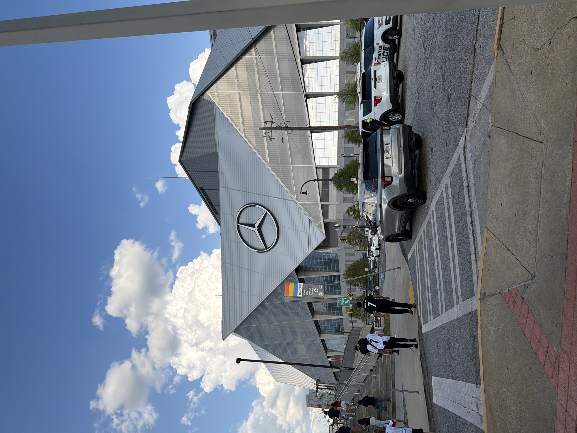 The image shows the exterior of Mercedes-Benz Stadium in Atlanta, Georgia. The stadium's distinctive architecture with the large Mercedes-Benz logo is prominent. Several vehicles, including a MARTA police car, are parked along the street adjacent to the stadium. Pedestrians are walking on the sidewalk and stairs leading up to the stadium entrance. The sky is blue with white clouds. A Marta transit sign is visible.