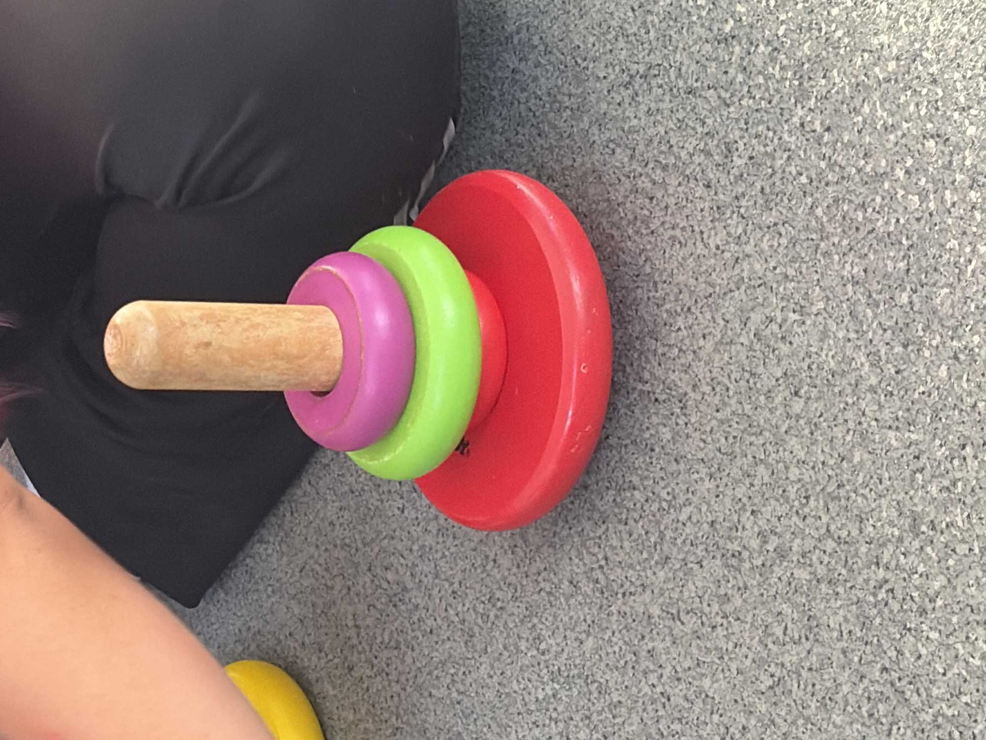 A wooden stacking toy with a red base, a green ring, a purple ring, and a wooden peg is centered on a gray speckled floor. To the left of the toy, a portion of a person's arm, clad in pale pink skin, is visible, and a bright yellow object, possibly a toy, is partially in frame. Behind the toy and person's arm, a person is wearing dark clothing. The scene appears to be indoors, likely in a home or daycare setting. The image does not provide any specific geographical information that would allow for identification of its location within Teplice, Czechia.