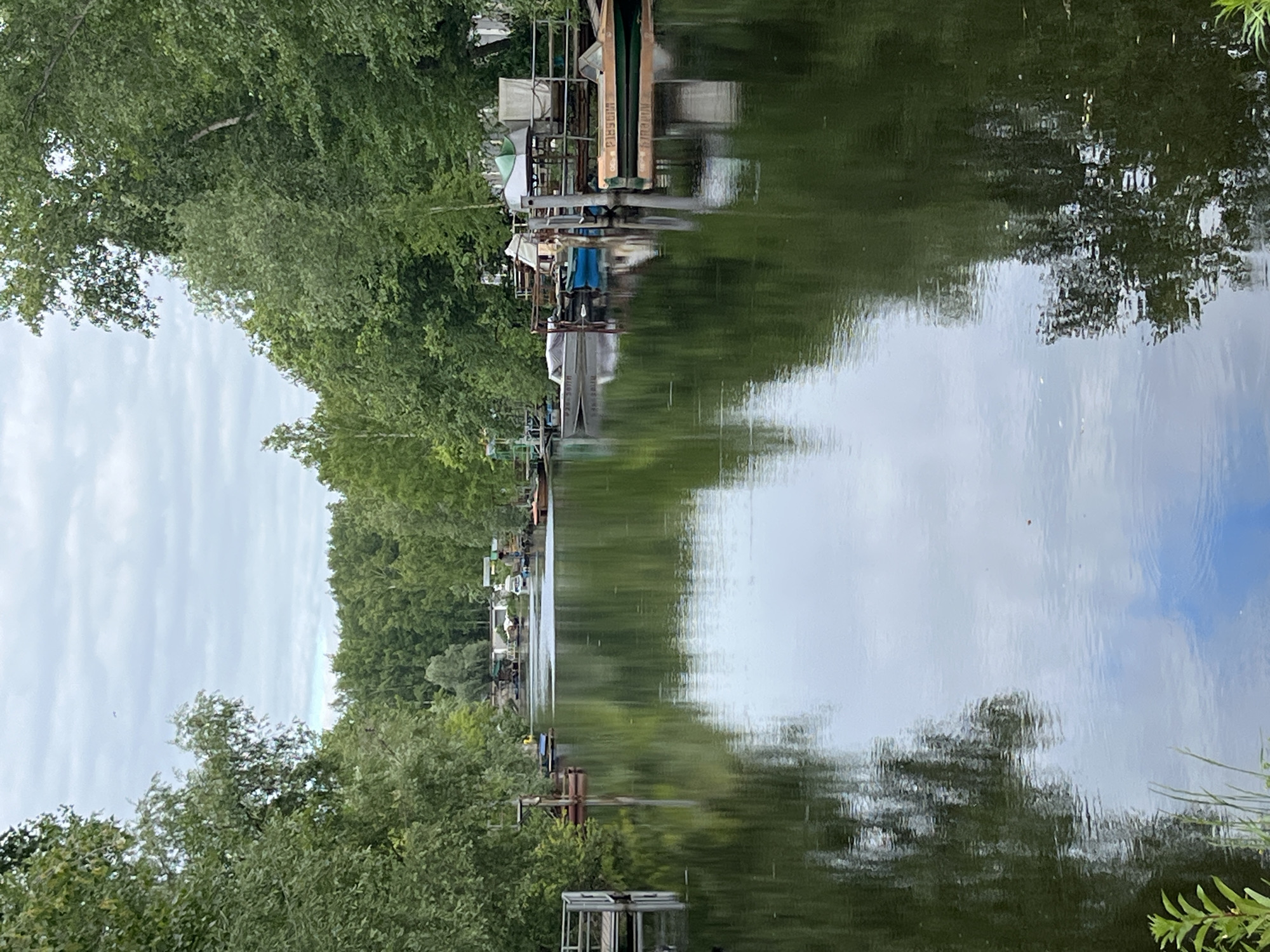 A calm, reflective river flows through a verdant, tree-lined channel in the city of Chaikovsky, Russia. The water mirrors the lush green foliage of the trees that densely flank both banks. Several boats are moored along the right bank, including a green and orange motorboat in the foreground, and various smaller vessels with covers and tarps. On the left bank, a wooden dock extends into the water, with another small platform structure further back. In the distance, more buildings and boats are visible, suggesting a residential or recreational area. The sky above is overcast with scattered clouds, and a single bird is visible in the upper left portion of the image. The scene depicts a peaceful, natural environment integrated with human activity.