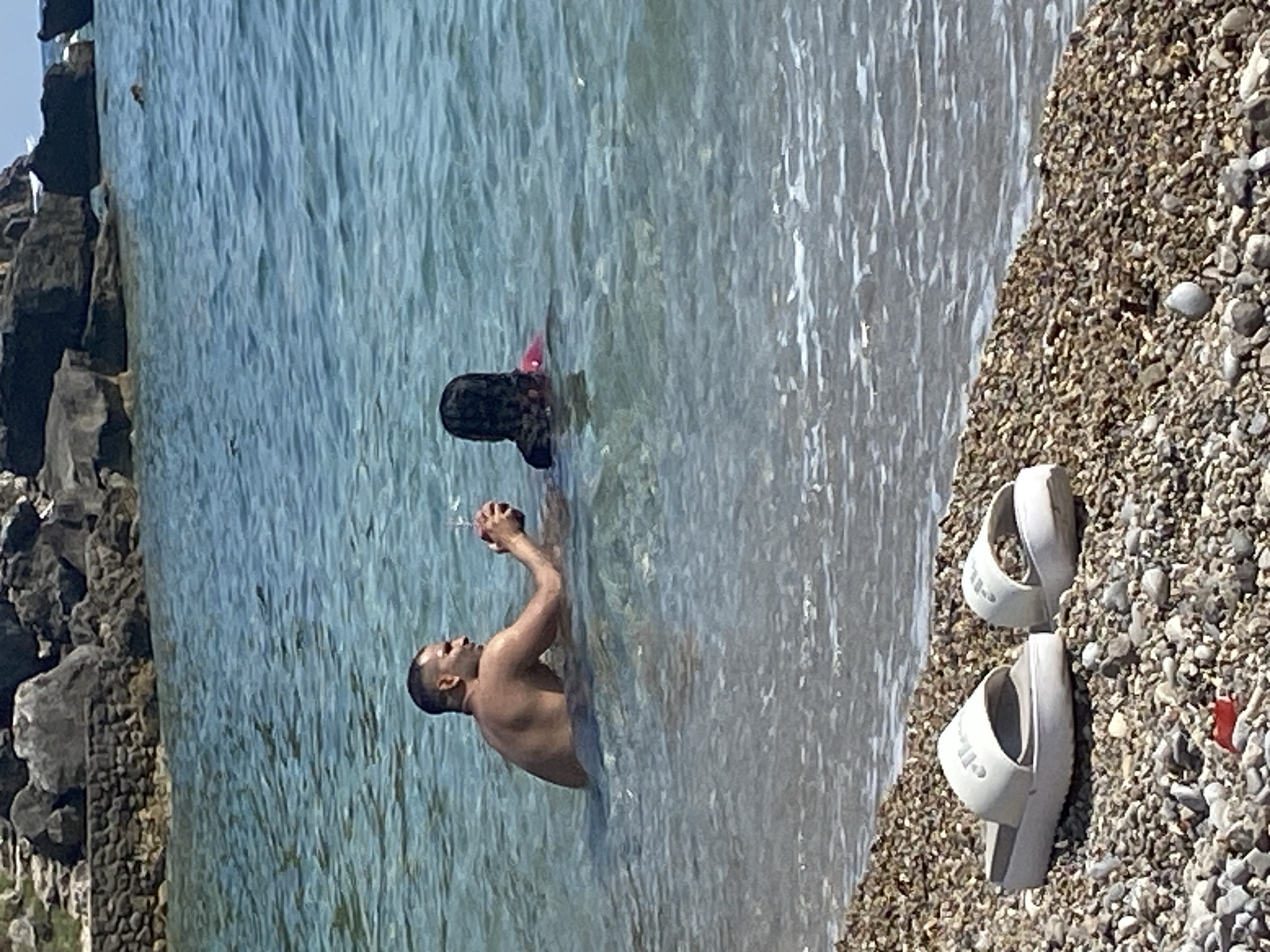 In Prijevor, Montenegro, two individuals are in the sea near a rocky shoreline. A man, partially submerged in the water, holds a drink. Beside him, another person, with dark hair and wearing a pink garment, is also in the water. In the foreground, two white sandals rest on a pebble beach. The water is clear and blue, with sunlight glinting off its surface. The background features large rocks and a stone wall.