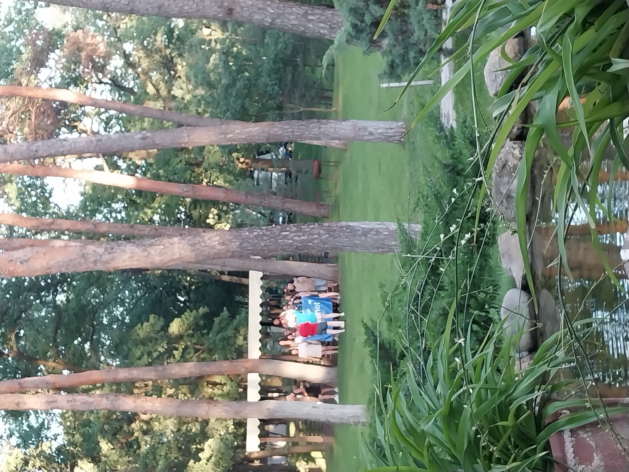 A group of people are gathered on a grassy area in a park-like setting with tall trees. Some individuals are holding a large blue banner. In the foreground, there is a body of water with aquatic plants. The scene appears to be outdoors during daylight hours. The location is Orlivshchyna, Ukraine.